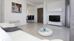 A modern living room with a white sofa, a flat-screen TV, a dining area with chairs, and a painting of the Eiffel Tower. - Sun Resort Nikolina (Bibinje)