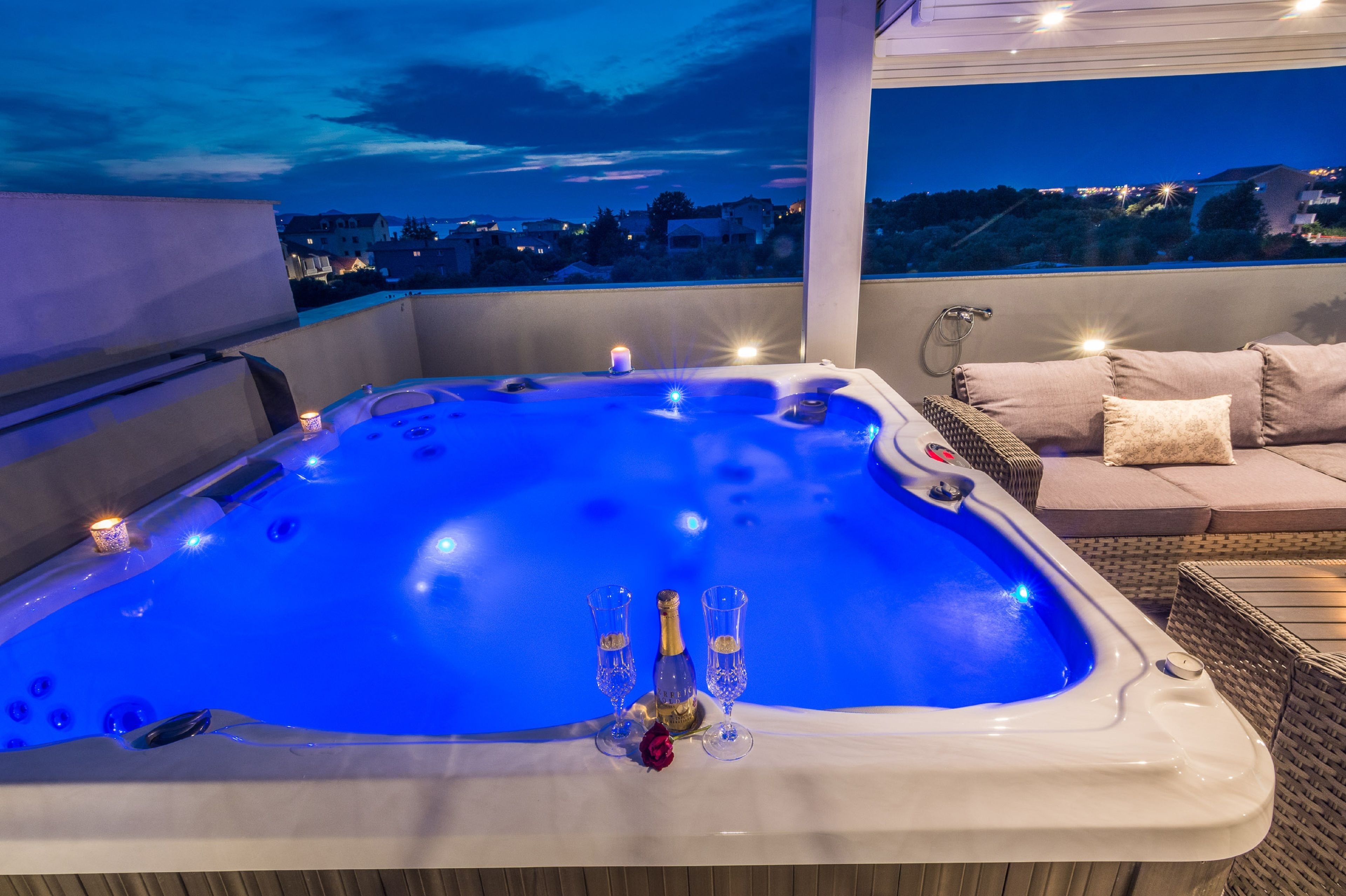 A hot tub with champagne glasses and a rose, surrounded by wicker furniture and a city view at dusk.