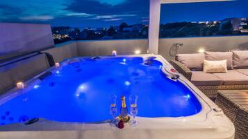 A hot tub with champagne glasses and a rose, surrounded by wicker furniture and a city view at dusk.