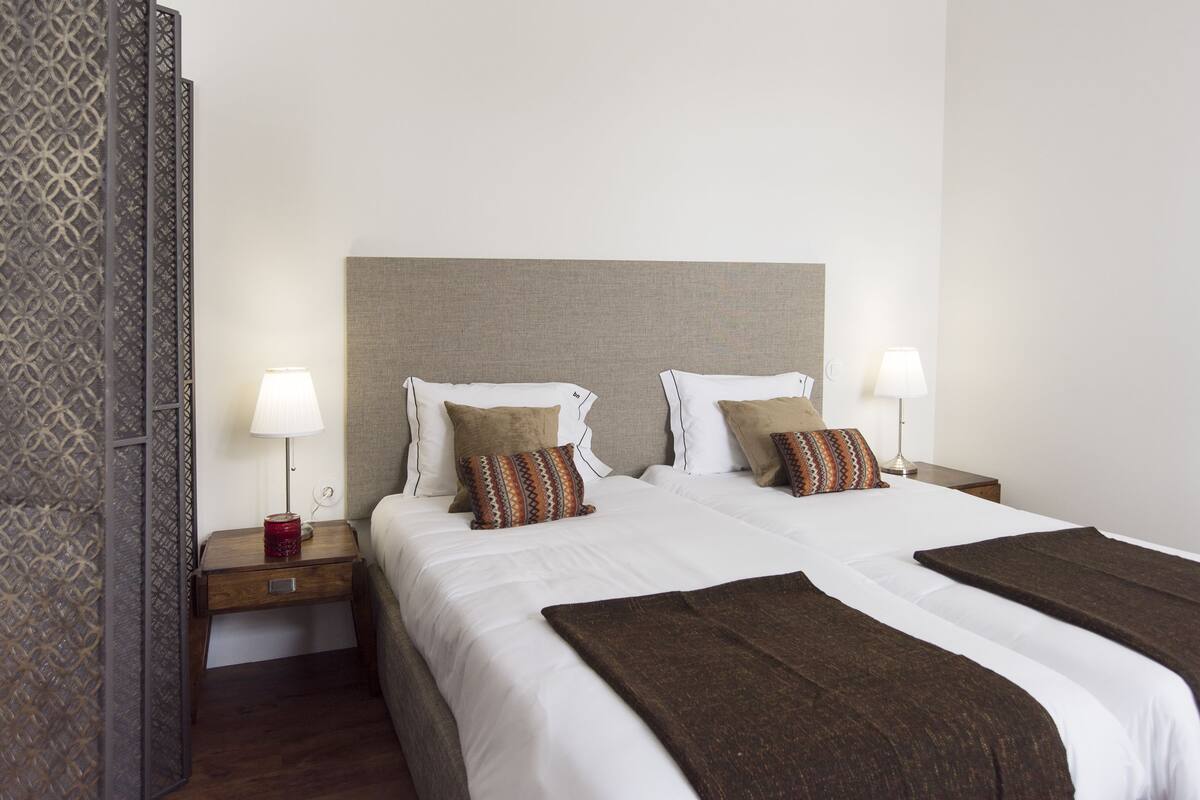 Habitación de hotel con una cama grande, dos mesitas de noche con lámparas, un separador decorativo y una paleta de colores neutros.