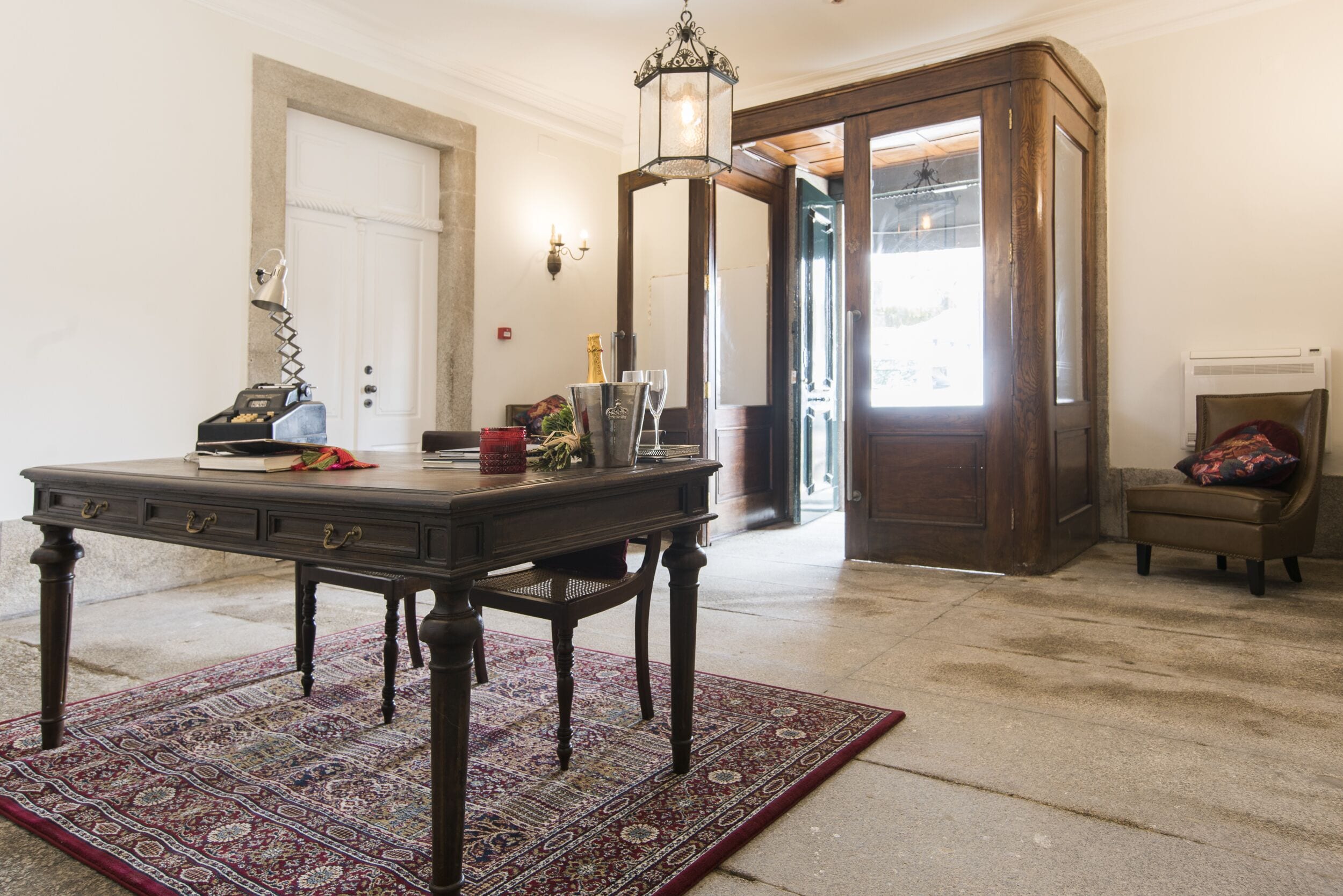 A wooden table with a rug, a lamp, and a chair in a room with a door and a window.