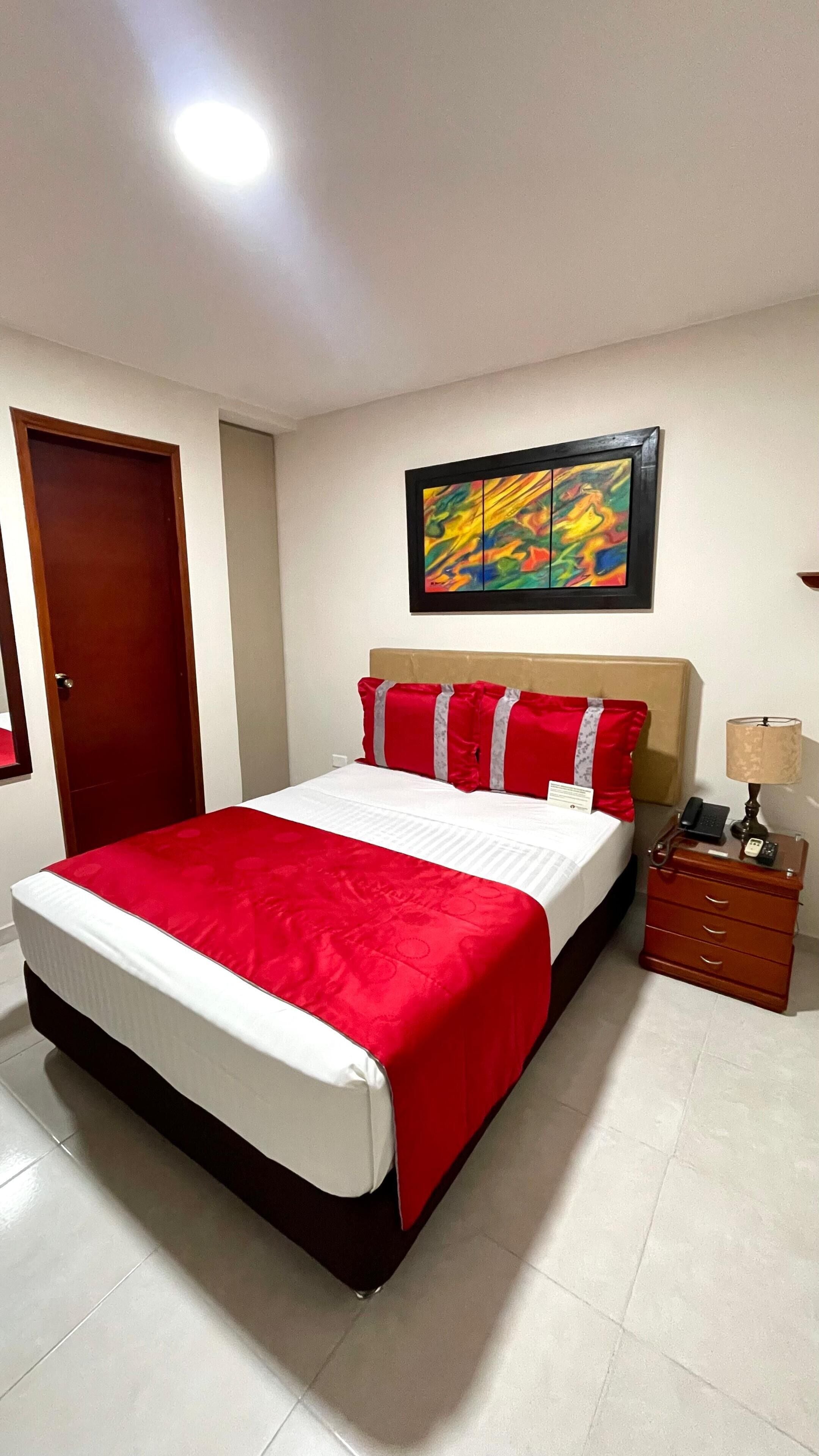 Une chambre d’hôtel avec un grand lit, une table de chevet en bois, une lampe et un tableau coloré accroché au mur.