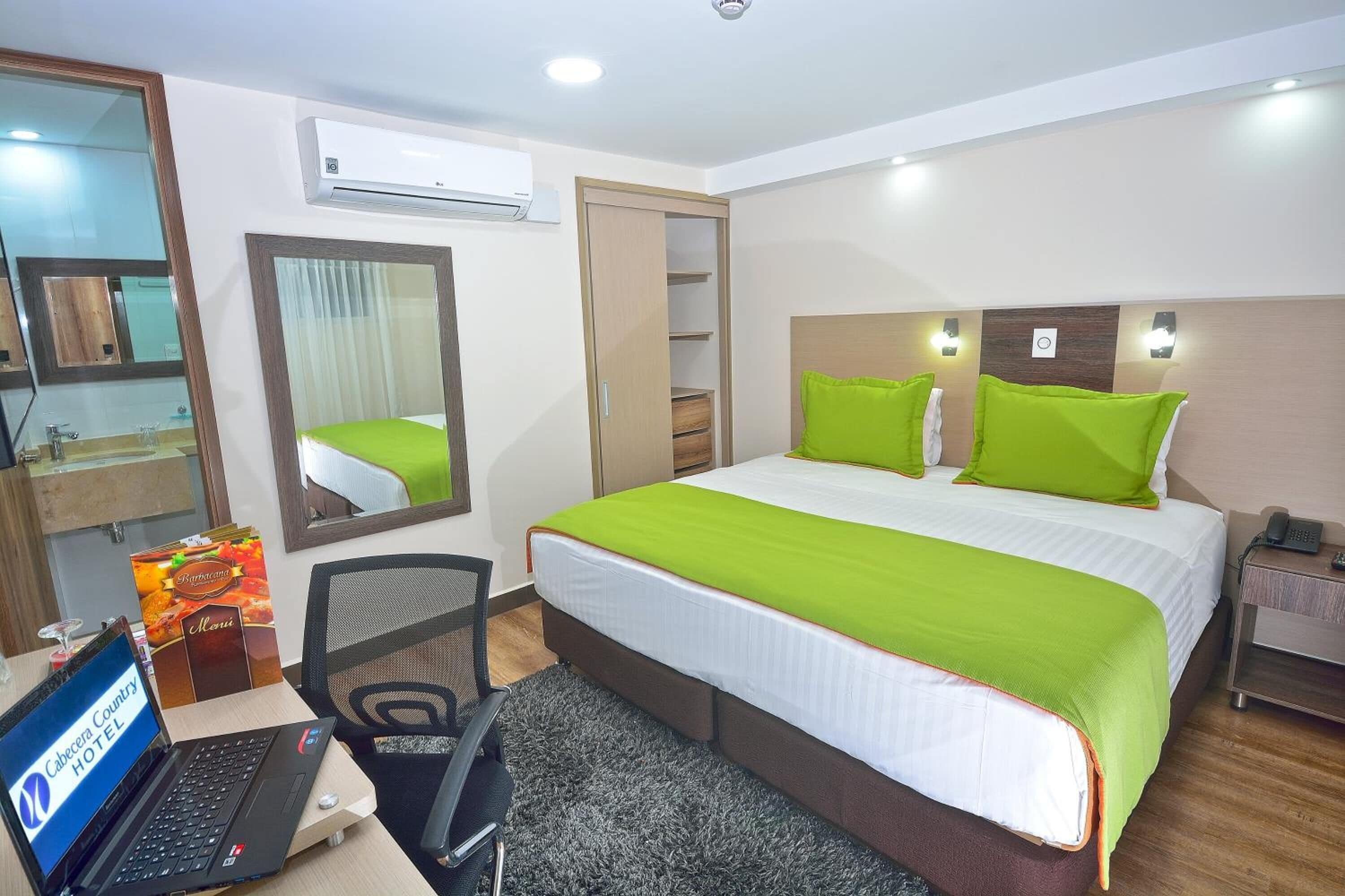 A hotel room with a bed, a desk with a laptop, a chair, a mirror, an air conditioning unit, and a nightstand with a phone.