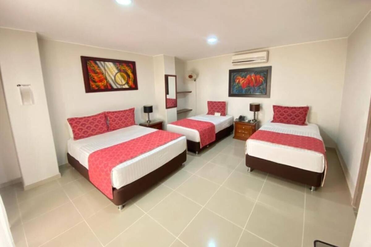 A hotel room with three beds, each with red bedding and white sheets. There are two wall-mounted air conditioning units, a painting on the wall, and a small table with a lamp.