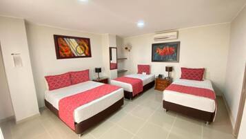 Habitación de hotel con tres camas, cada una con ropa de cama roja y sábanas blancas. Hay dos unidades de aire acondicionado fijadas a la pared, un cuadro y una pequeña mesa con una lámpara.