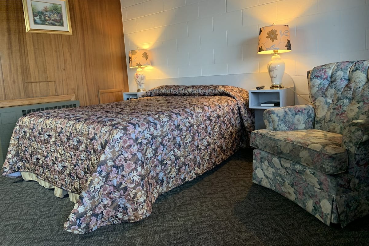 A bedroom with a floral bedspread, a wooden headboard, two bedside lamps, a floral armchair, and a small bedside table.