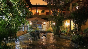 A courtyard with a table and chairs under an umbrella, surrounded by lush greenery and a stone wall.