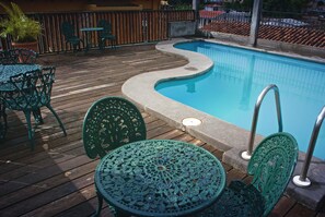 A pool with a curved edge, surrounded by a wooden deck with green metal chairs and tables.