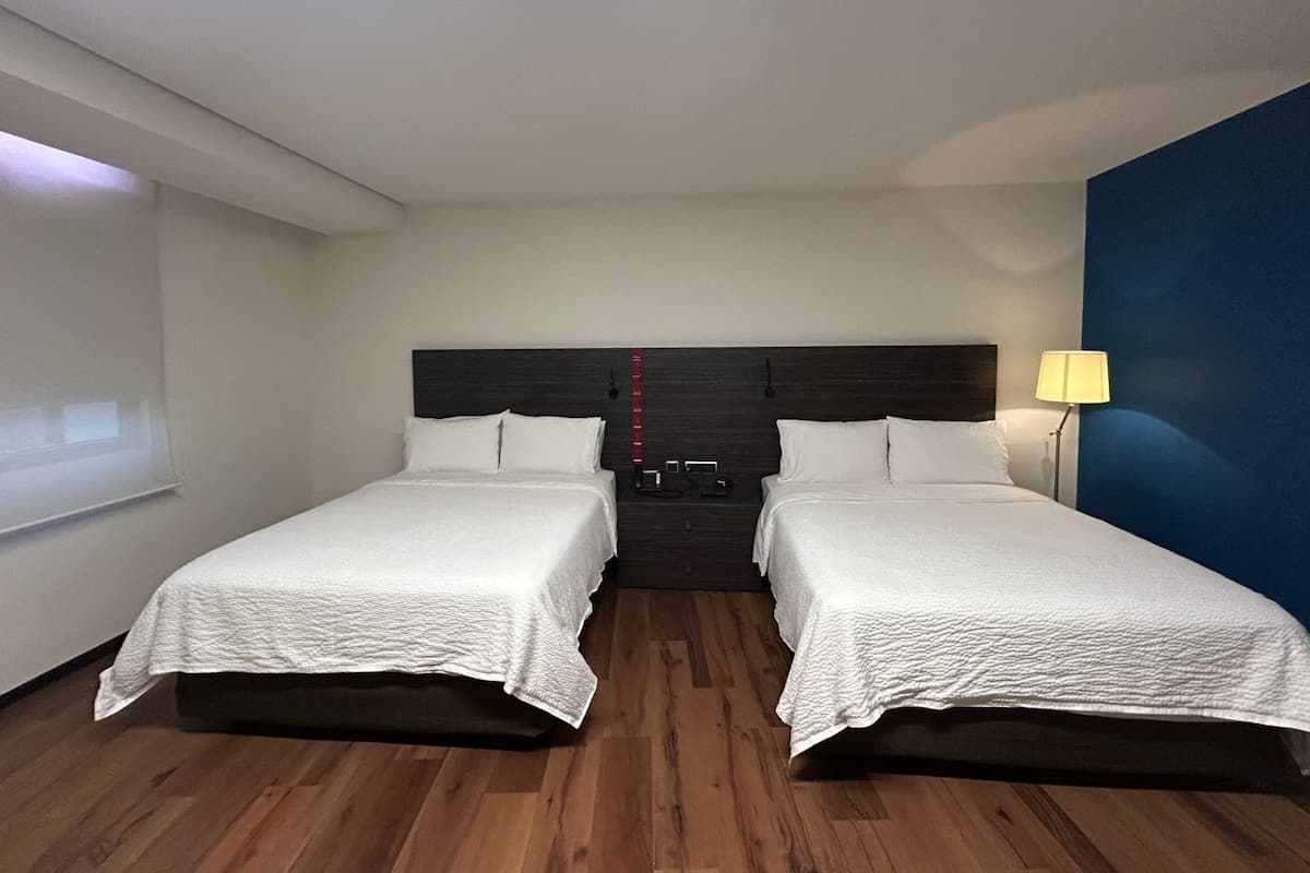 A hotel room with two single beds, a dark headboard, a bedside lamp, and a projector screen on the left wall.