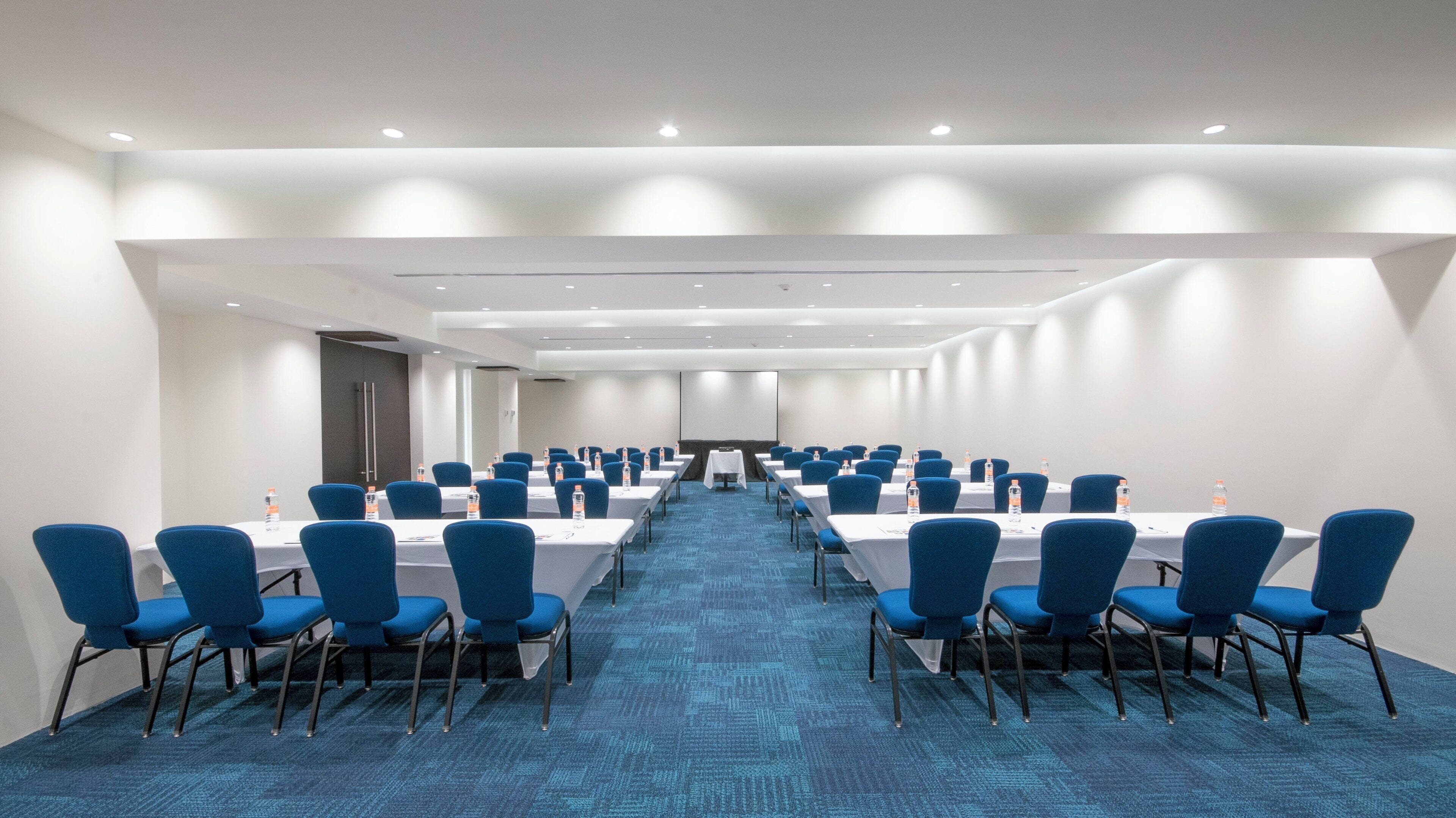 Un salón de conferencias con filas de mesas y sillas, equipado con botellas de agua.