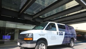 Una camioneta blanca con la leyenda "SERVICIO GRATUITO FREE SERVICE" y el logotipo de "Holiday Inn Express" estacionada frente a un edificio de fachada de vidrio.