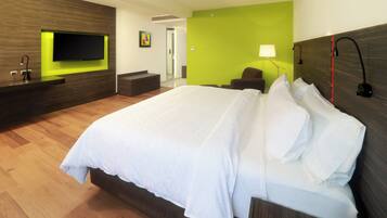 A hotel room with a large bed, a flat-screen TV mounted on a wooden panel, a green accent wall, a desk with a chair, and a bedside lamp.