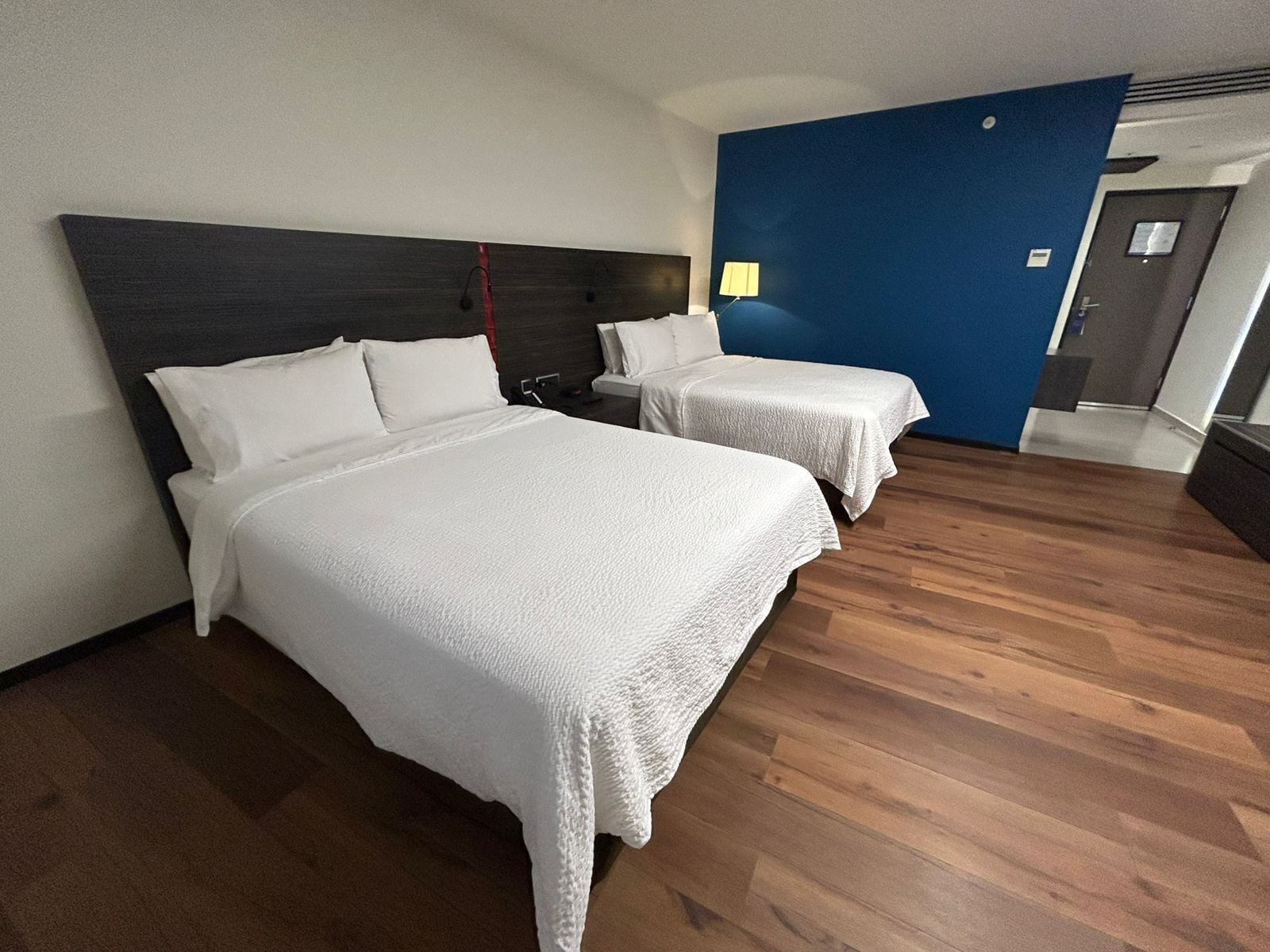 Ein Hotelzimmer mit zwei Betten, einem dunklen Kopfteil, einer blauen Akzentwand und Holzboden.