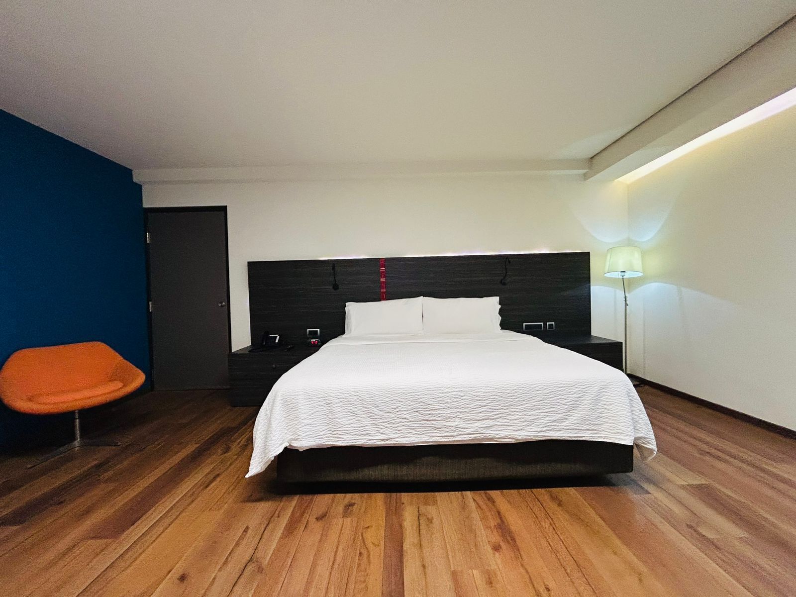 A modern hotel room with a large bed, a nightstand, a chair, and a bedside table.