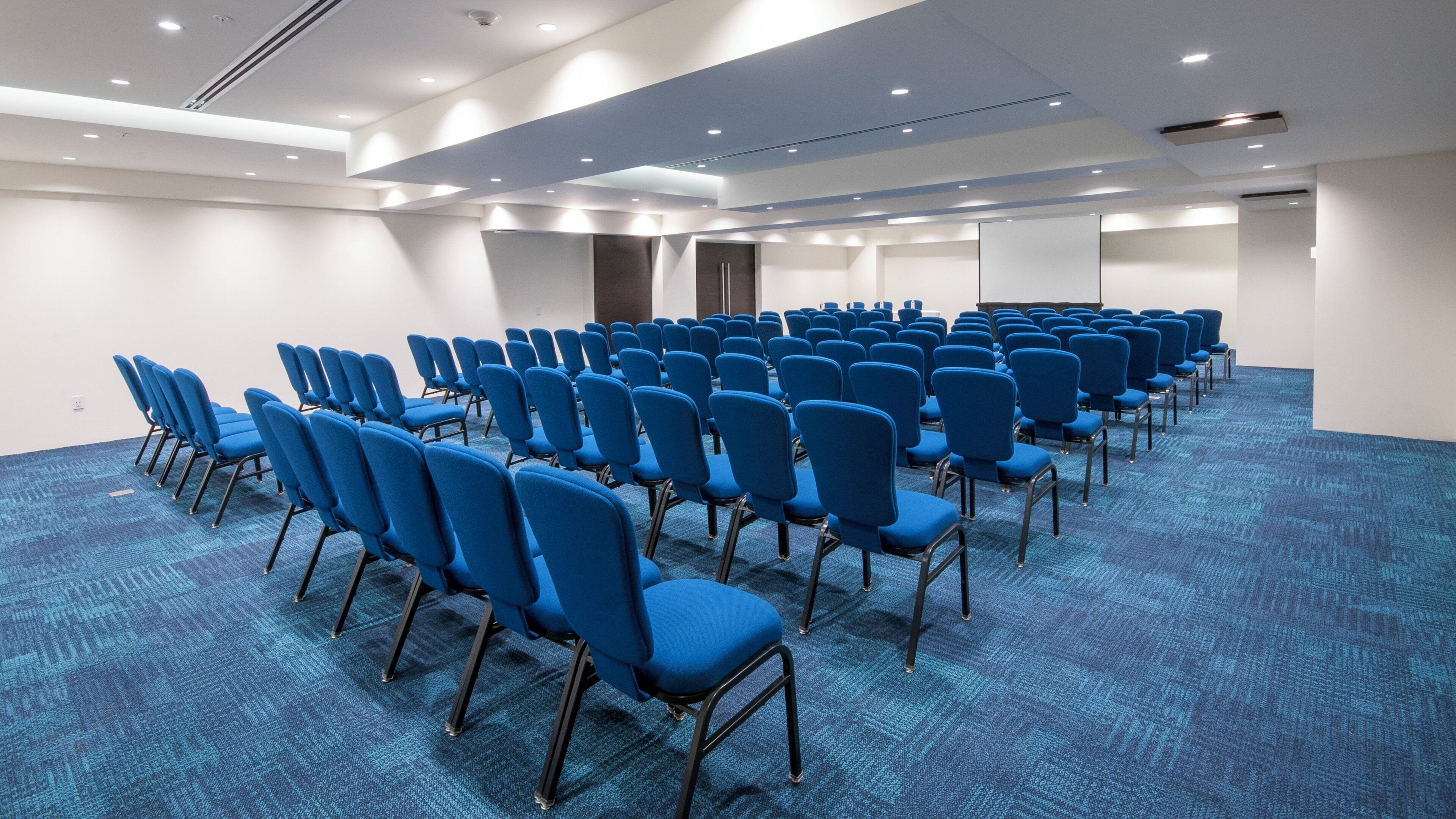 A conference room with rows of blue chairs, a carpeted floor, and a white ceiling with recessed lighting.