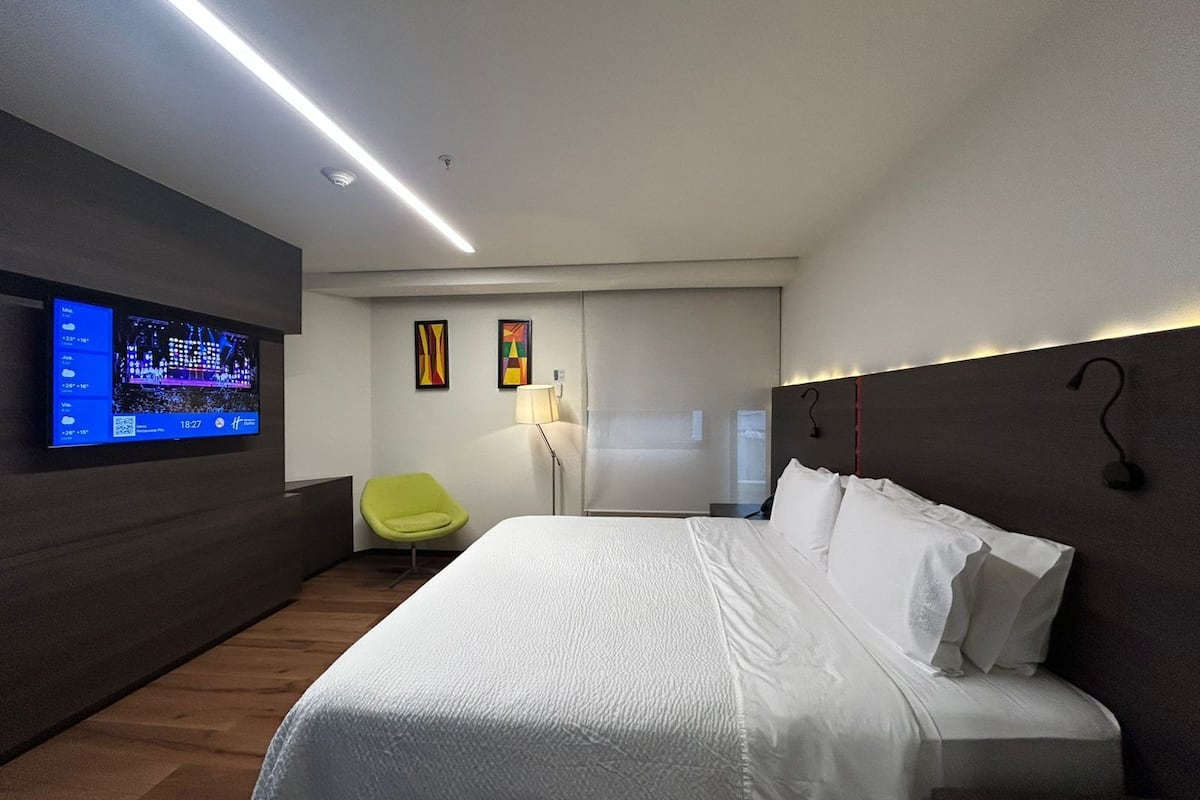 A modern hotel room with a large bed, a flat-screen TV mounted on the wall, a bright green chair, and a bedside table with a lamp.