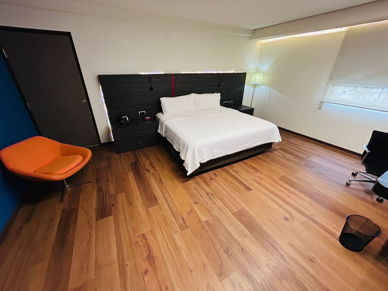 A modern bedroom with a wooden floor, a bed with white bedding, a desk, and an orange chair.