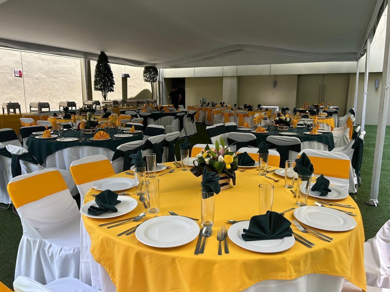 Un salón de banquetes con mesas redondas, cada una dispuesta con platos blancos, cubiertos y servilletas, y cintas amarillas en las sillas.