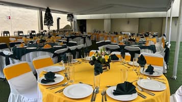 Une salle de banquet avec des tables rondes, chacune agrémentée d’assiettes blanches, de couverts et de serviettes, et des rubans jaunes sur les chaises.