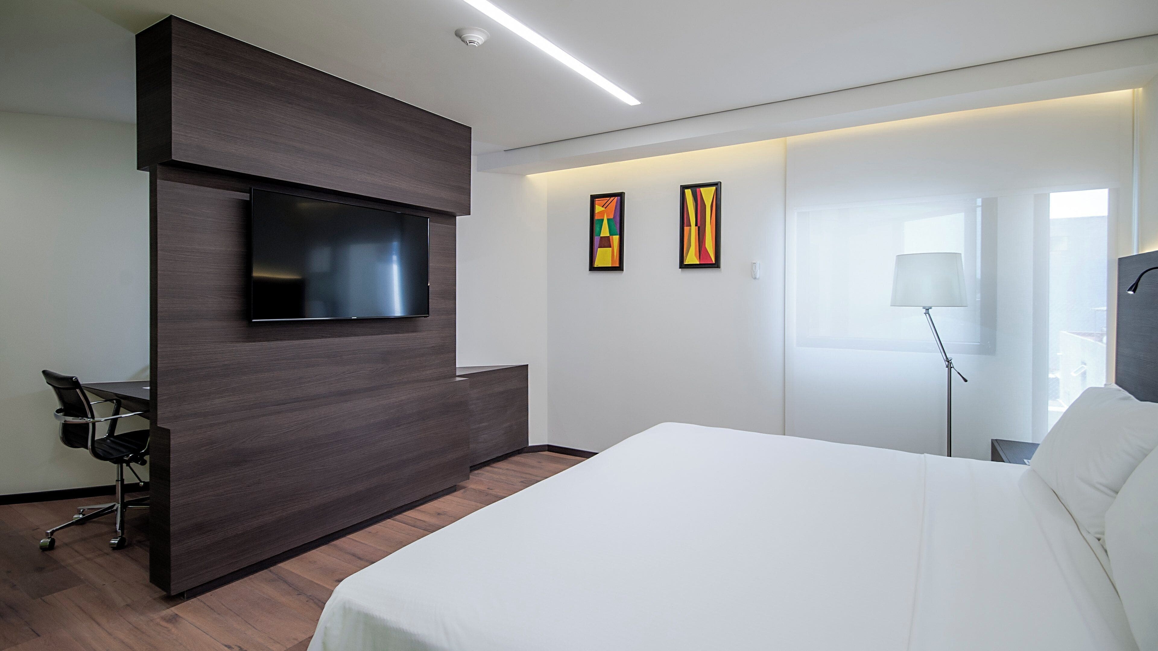 Quarto de hotel moderno com uma cama grande, uma TV de tela plana fixada em um painel de madeira escura, uma mesa com uma cadeira e duas pinturas abstratas na parede.