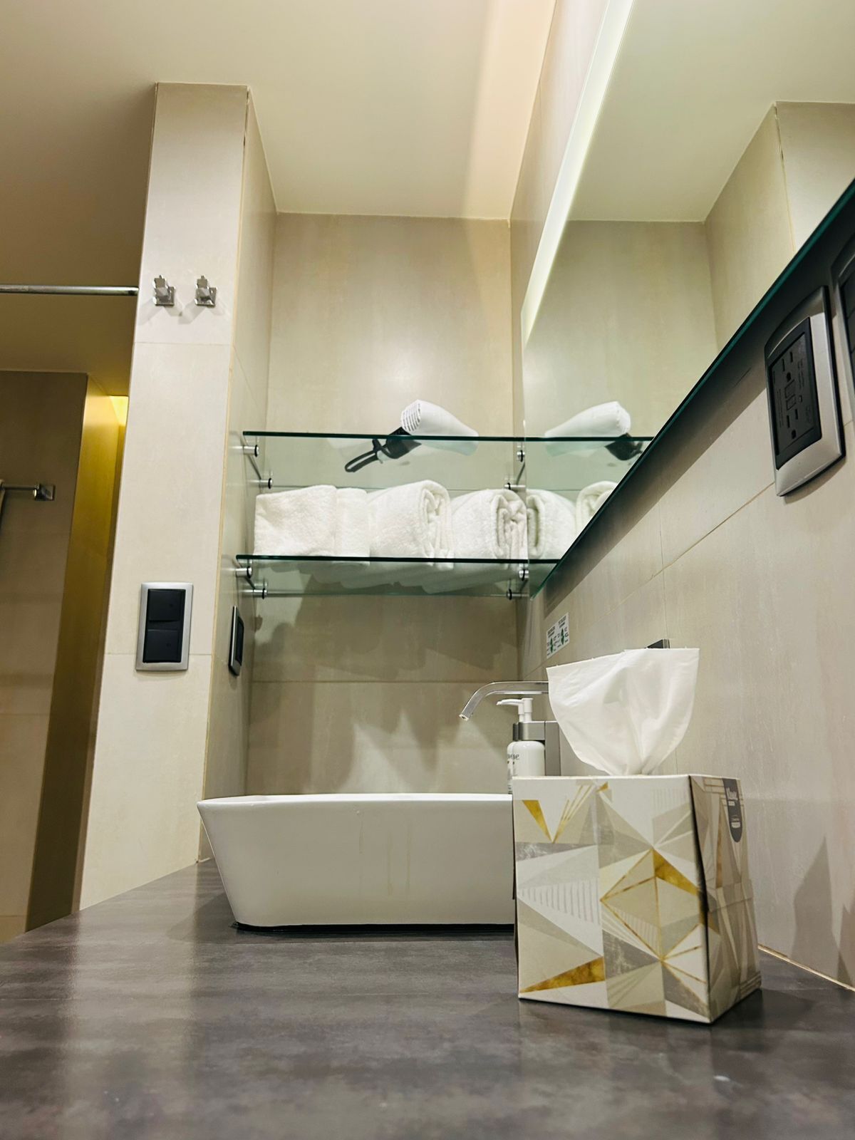 A modern bathroom with a bathtub, a glass shelf with towels, a hand dryer, and a tissue box.