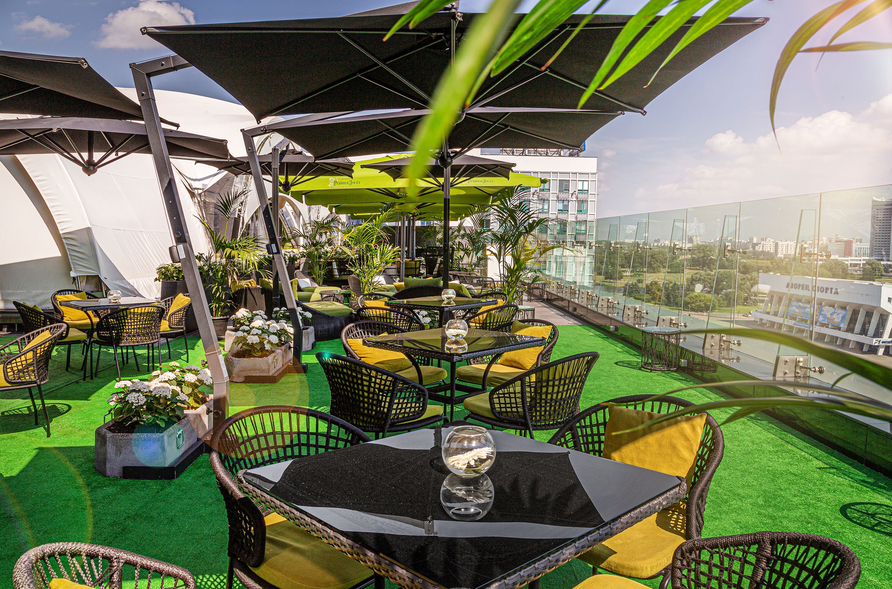 An outdoor seating area with tables, chairs, and umbrellas, surrounded by greenery and a city view.