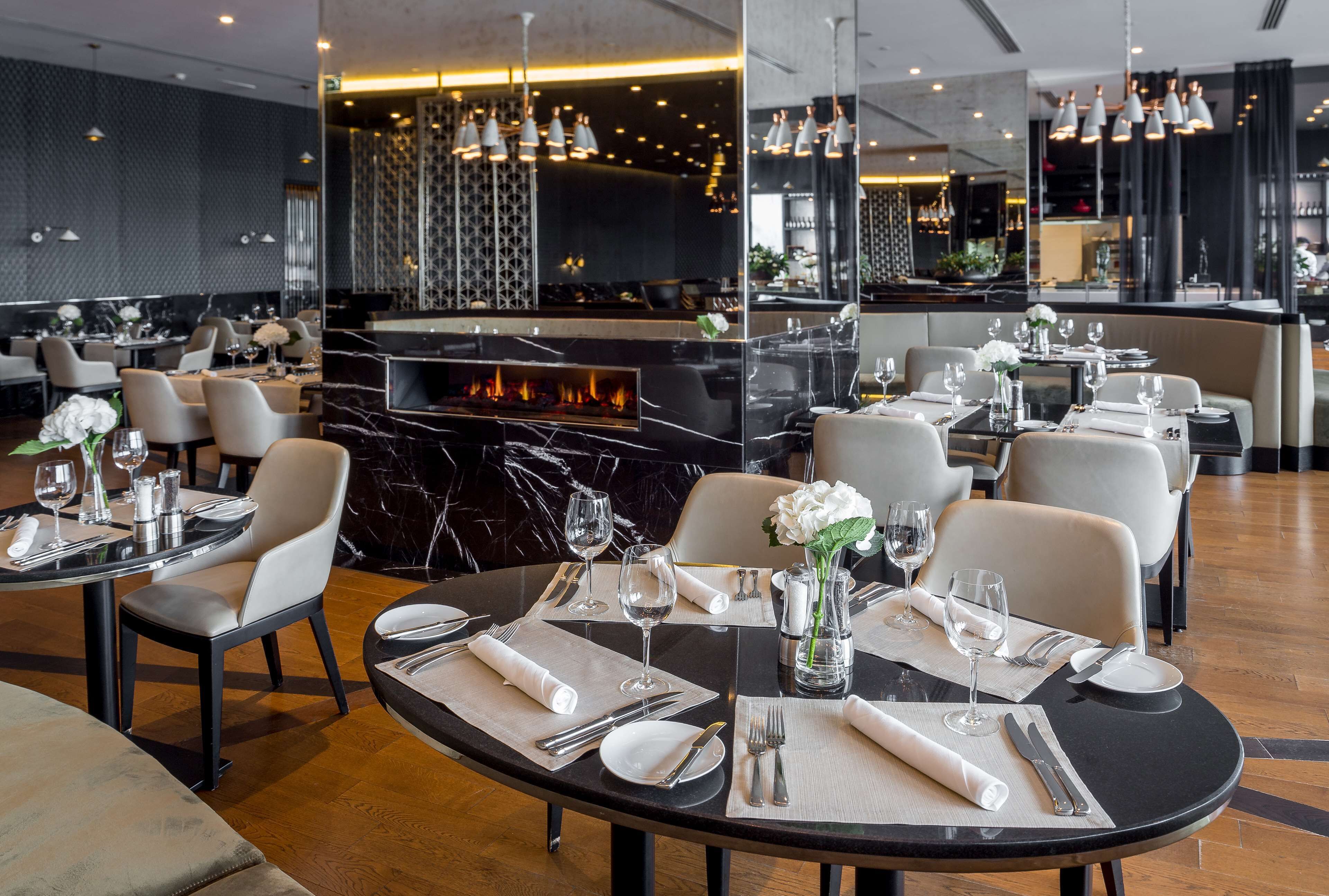 Een modern restaurant met ronde tafels, witte stoelen en een centraal geplaatst haardvuur.