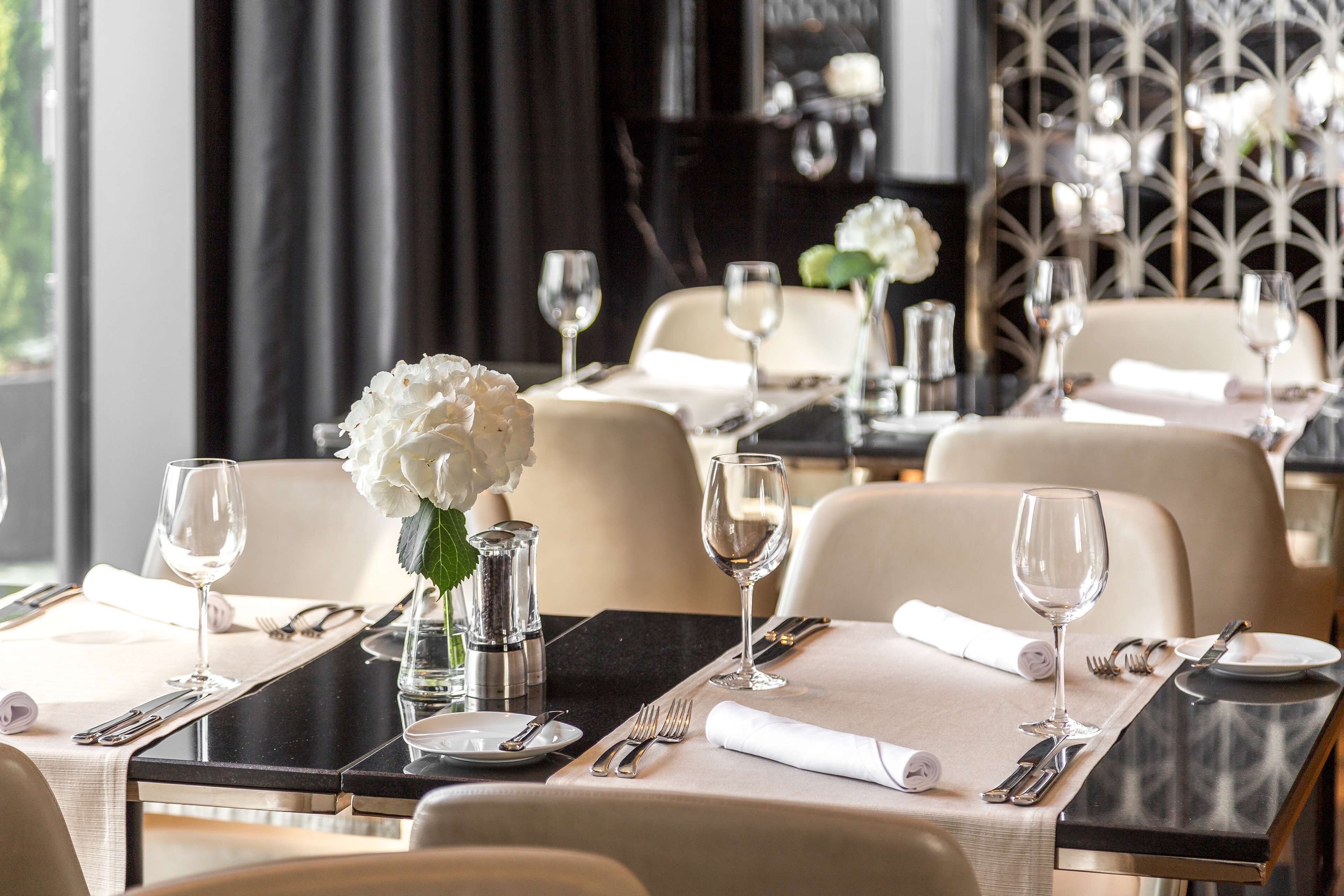 A dining area with tables set for two, each with a vase of white flowers, silverware, and wine glasses.