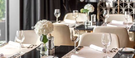 A dining area with tables set for two, each with a vase of white flowers, silverware, and wine glasses.