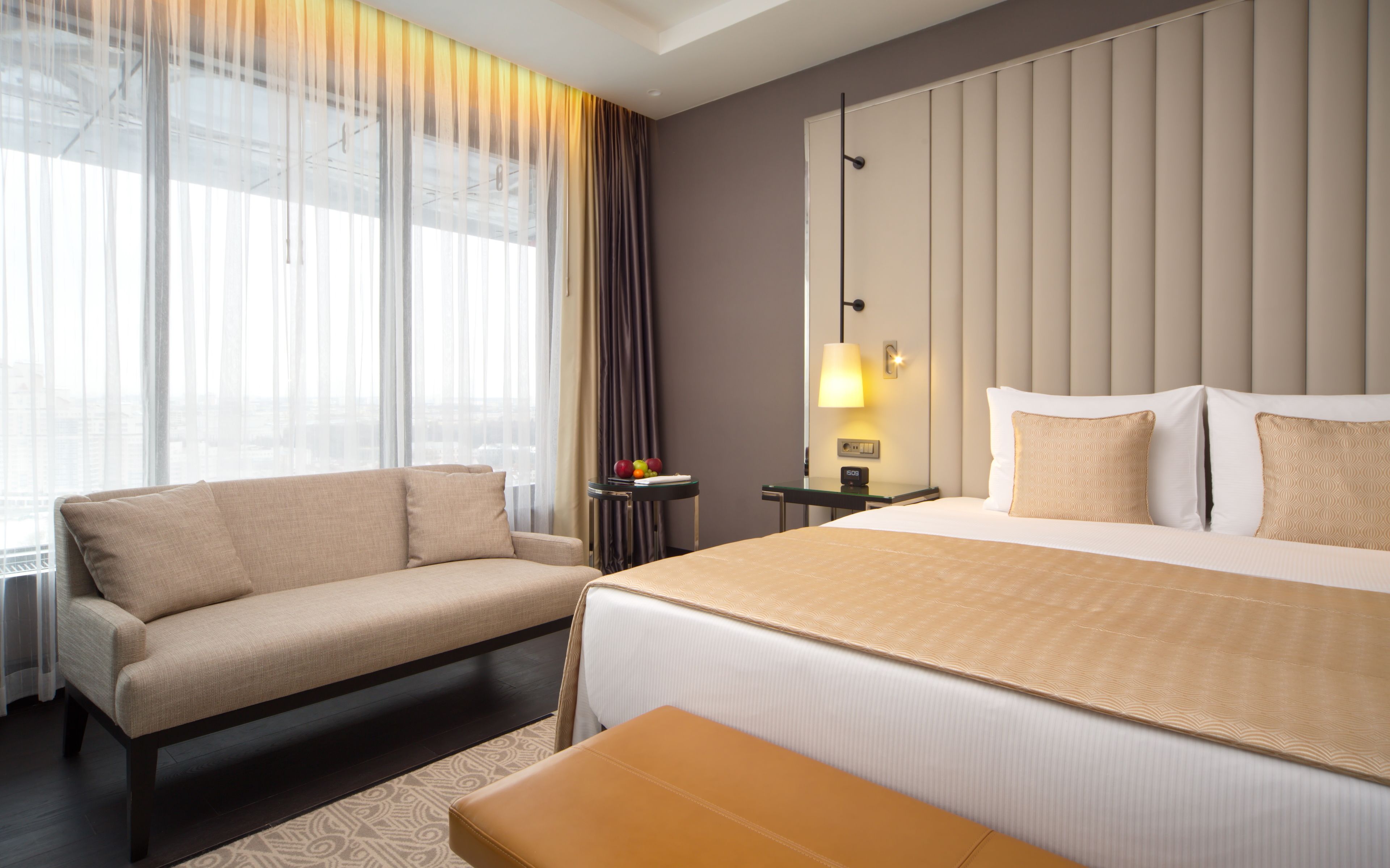 A hotel room with a bed, a sofa, a small table with fruit, a lamp, and a view of the city through large windows.