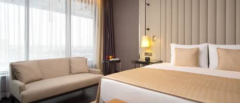 A hotel room with a bed, a sofa, a small table with fruit, a lamp, and a view of the city through large windows.