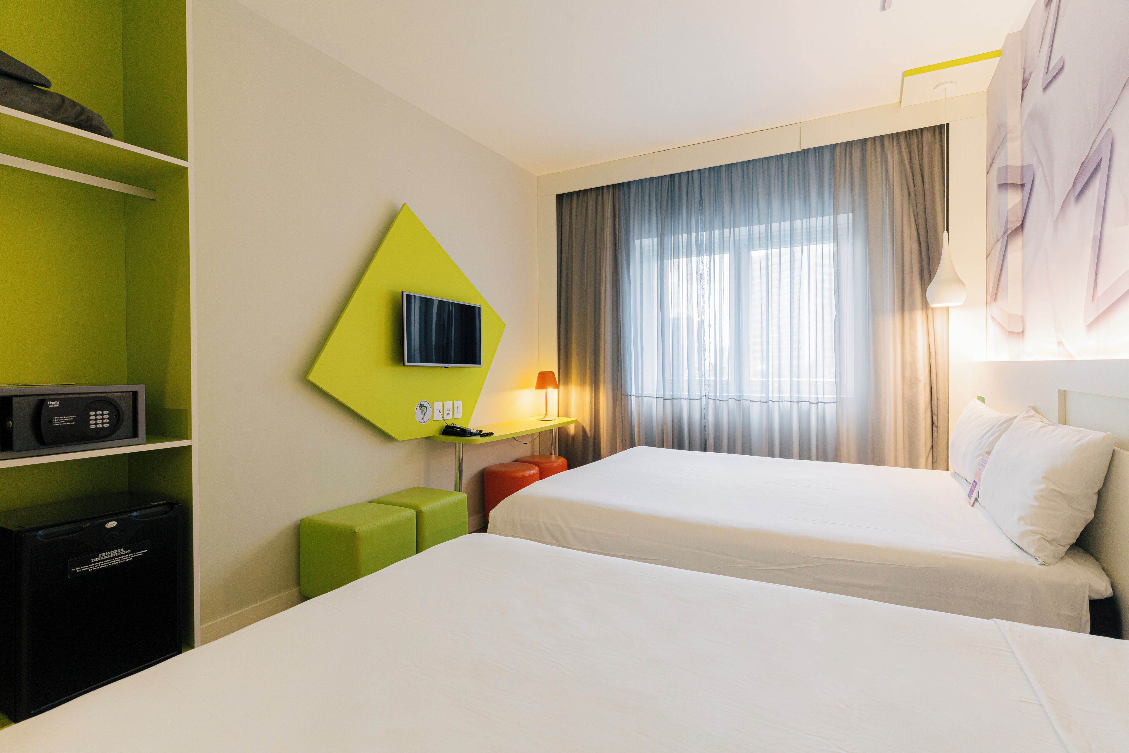 A hotel room with two beds, a TV, a desk with a lamp, and a green accent wall.