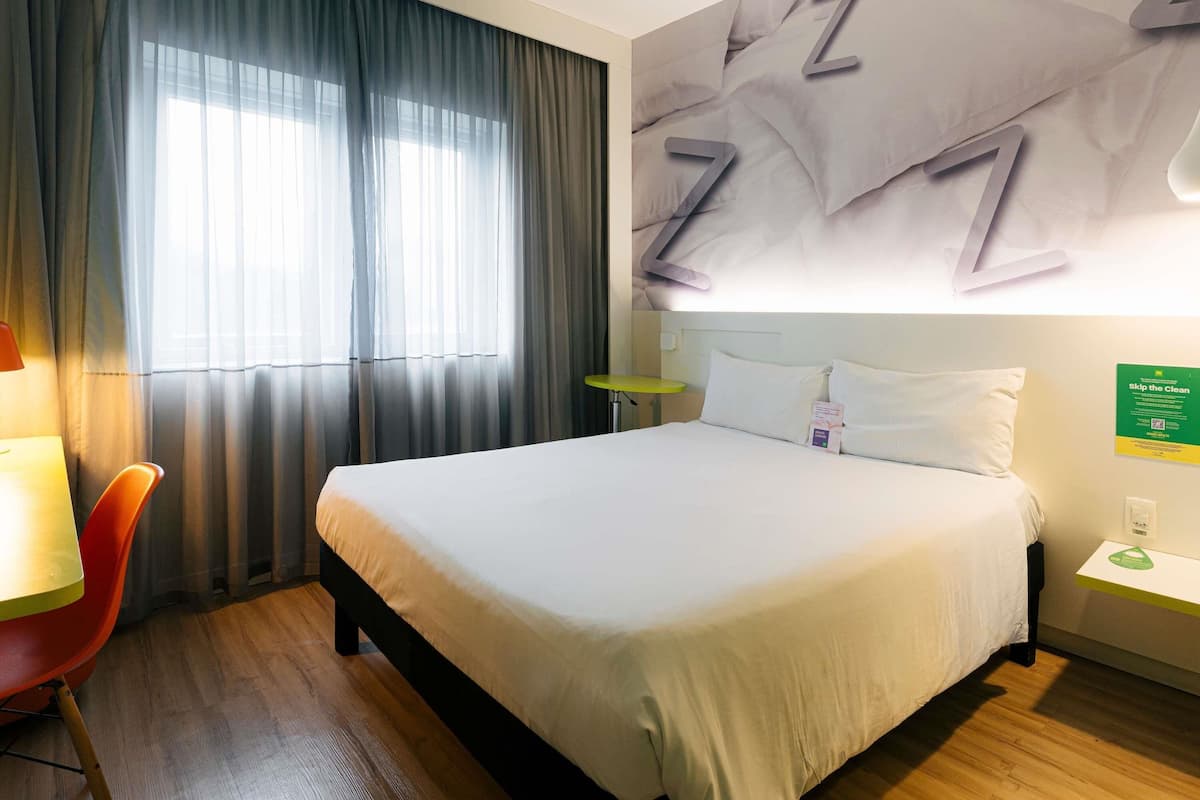 Hotelkamer met een bed, bureau, stoel, raam met gordijnen en een muur met een groot abstract ontwerp.