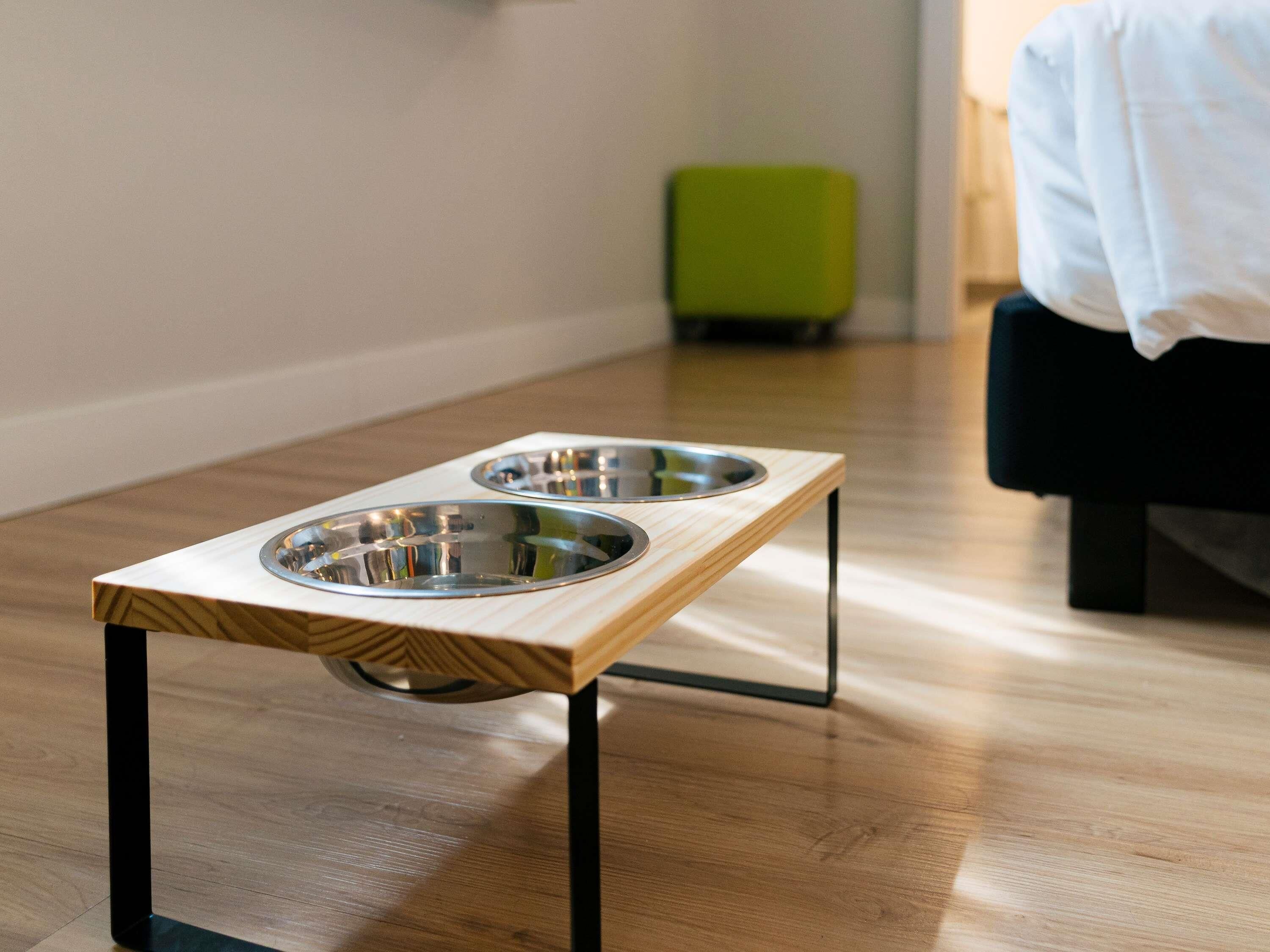 A wooden pet feeding station with two metal bowls on a hardwood floor.