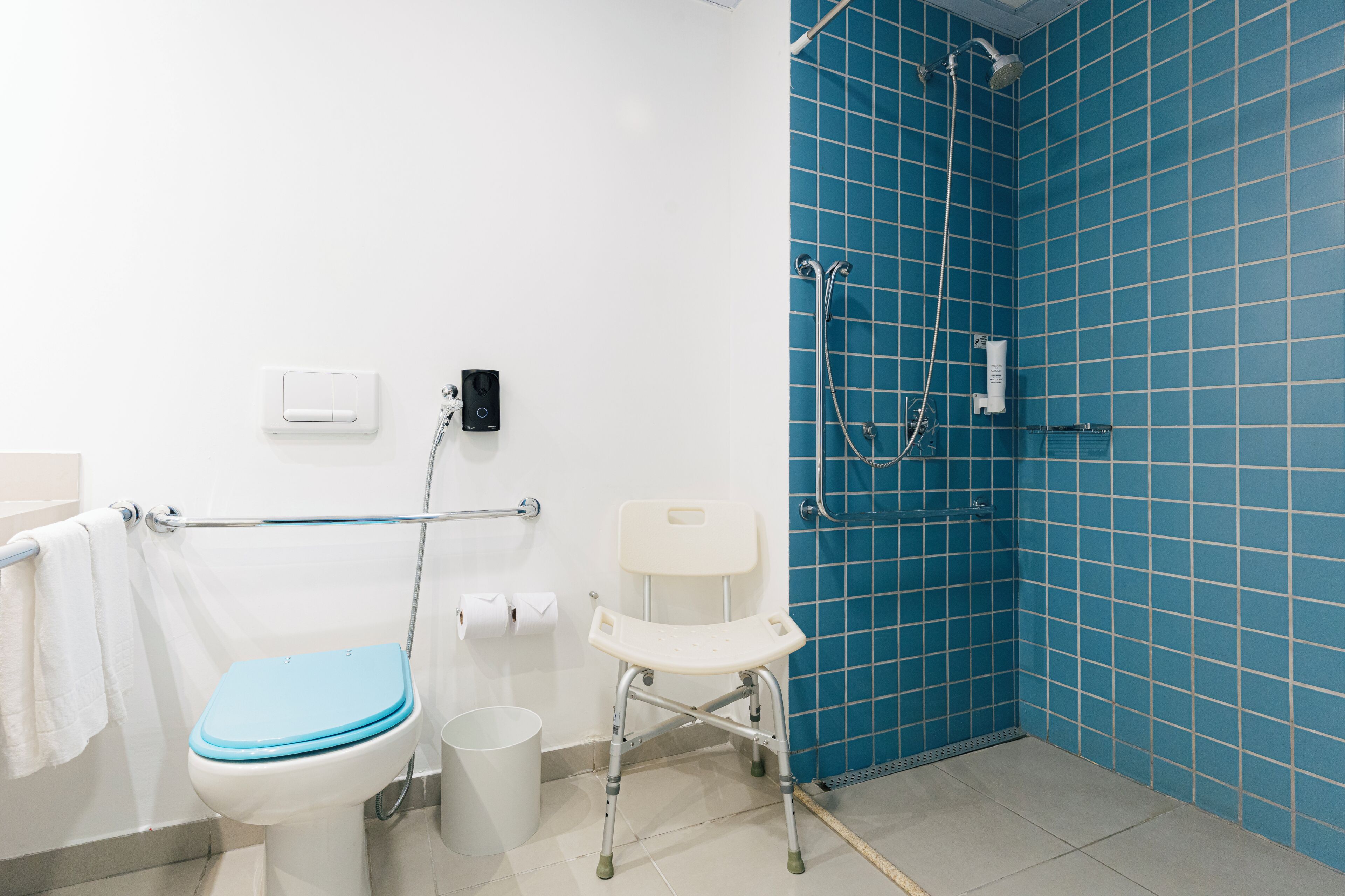 Casa de banho com sanita branca, chuveiro azul e barra de apoio branca.