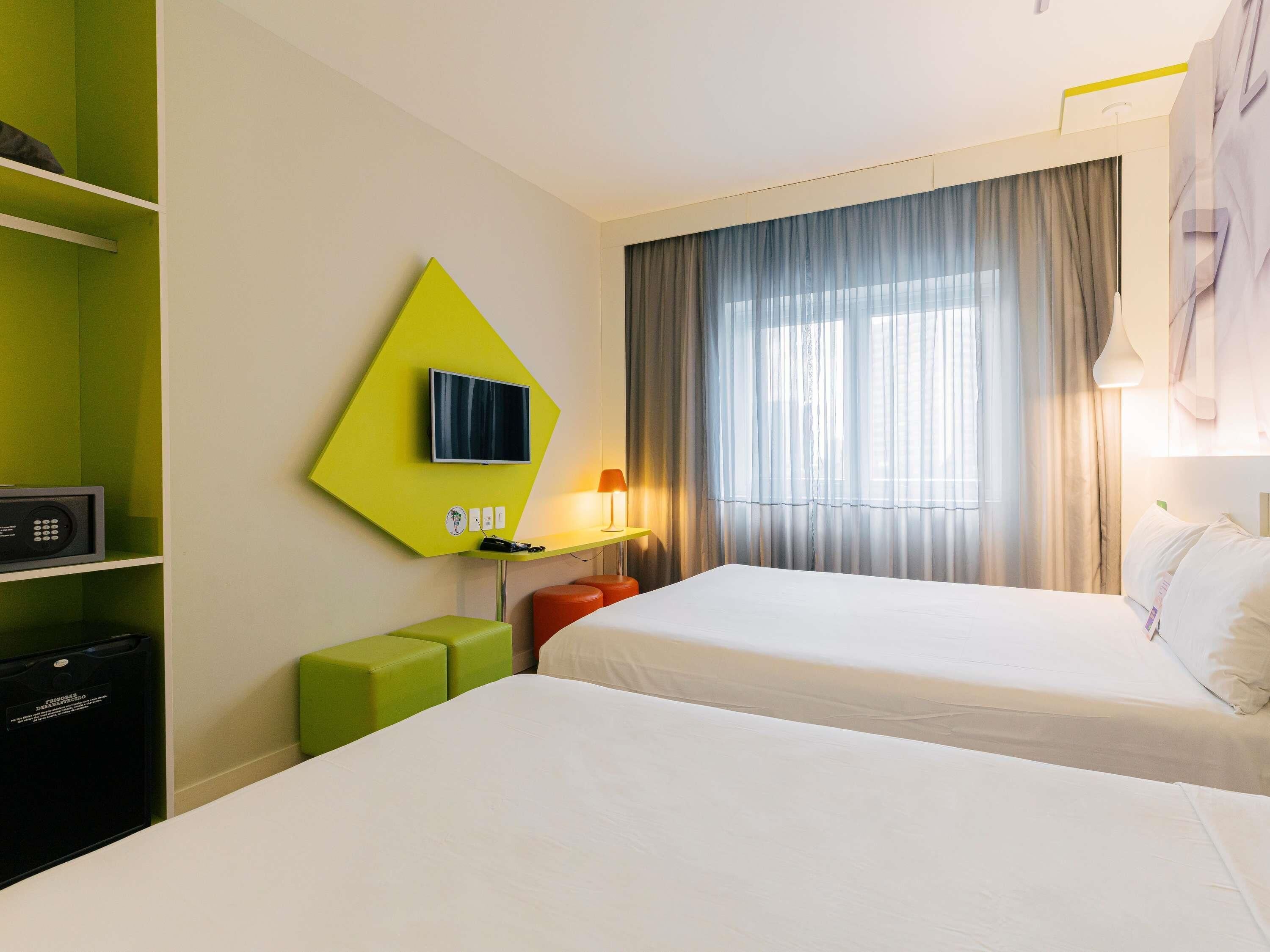 Une chambre d’hôtel avec deux lits, un téléviseur fixé sur un mur vert, un bureau avec une lampe et une fenêtre avec des rideaux.