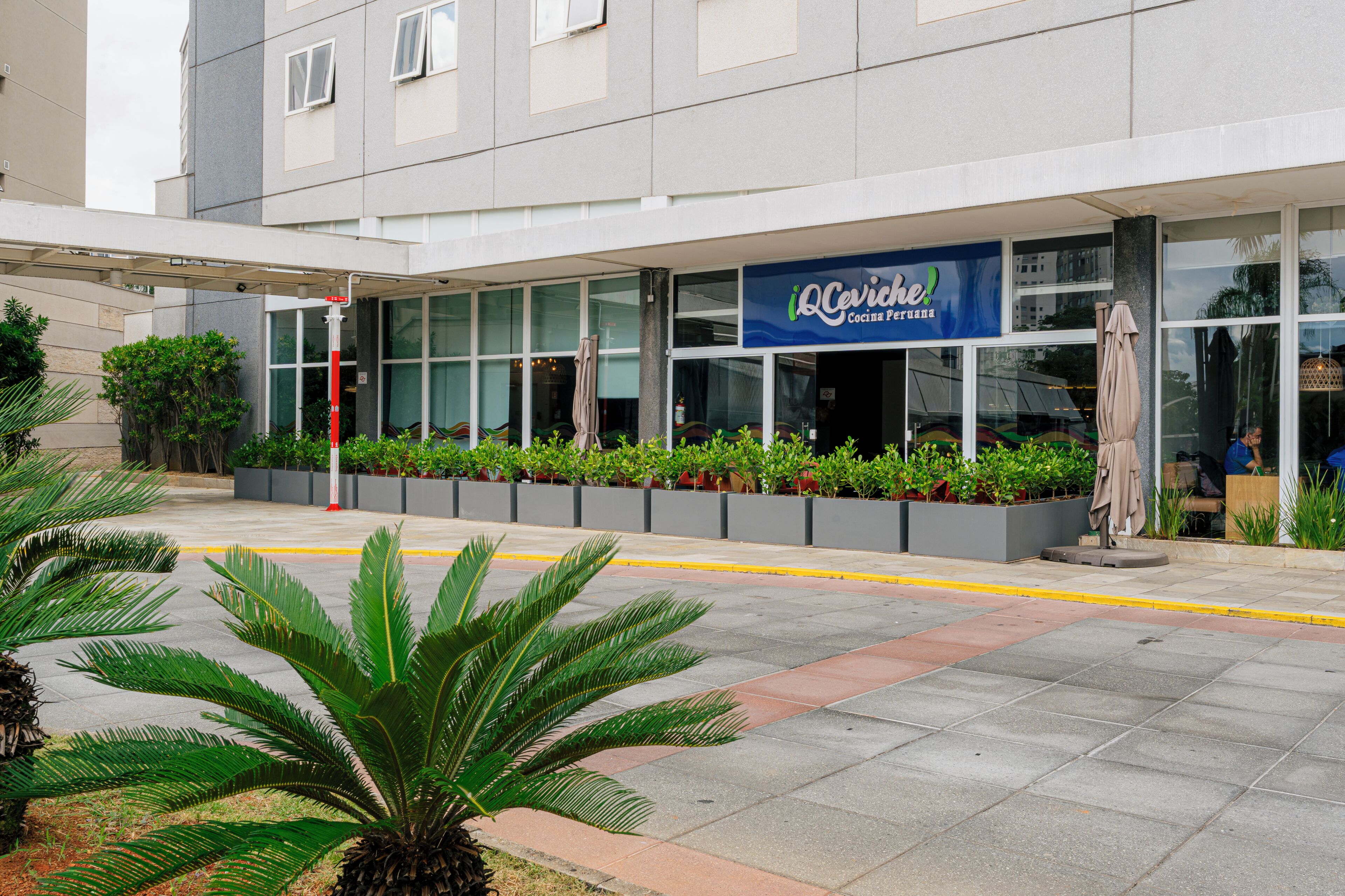 A modern building with a sign reading 'Cocina Peruana' and a covered outdoor area with planters and a palm tree.