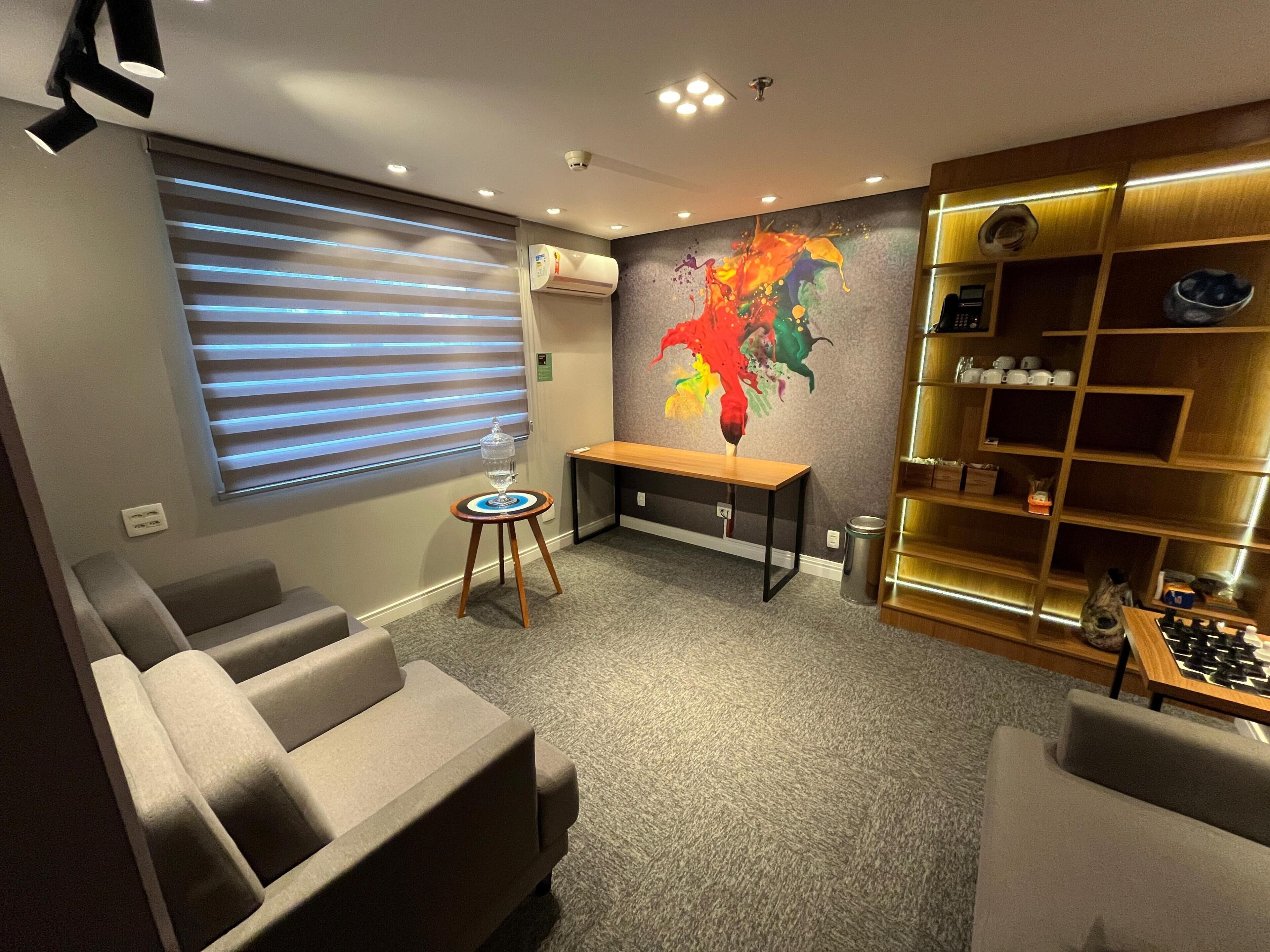 A modern living room with a sofa, a small table, a colorful abstract painting, a bookshelf, and a window with blinds.