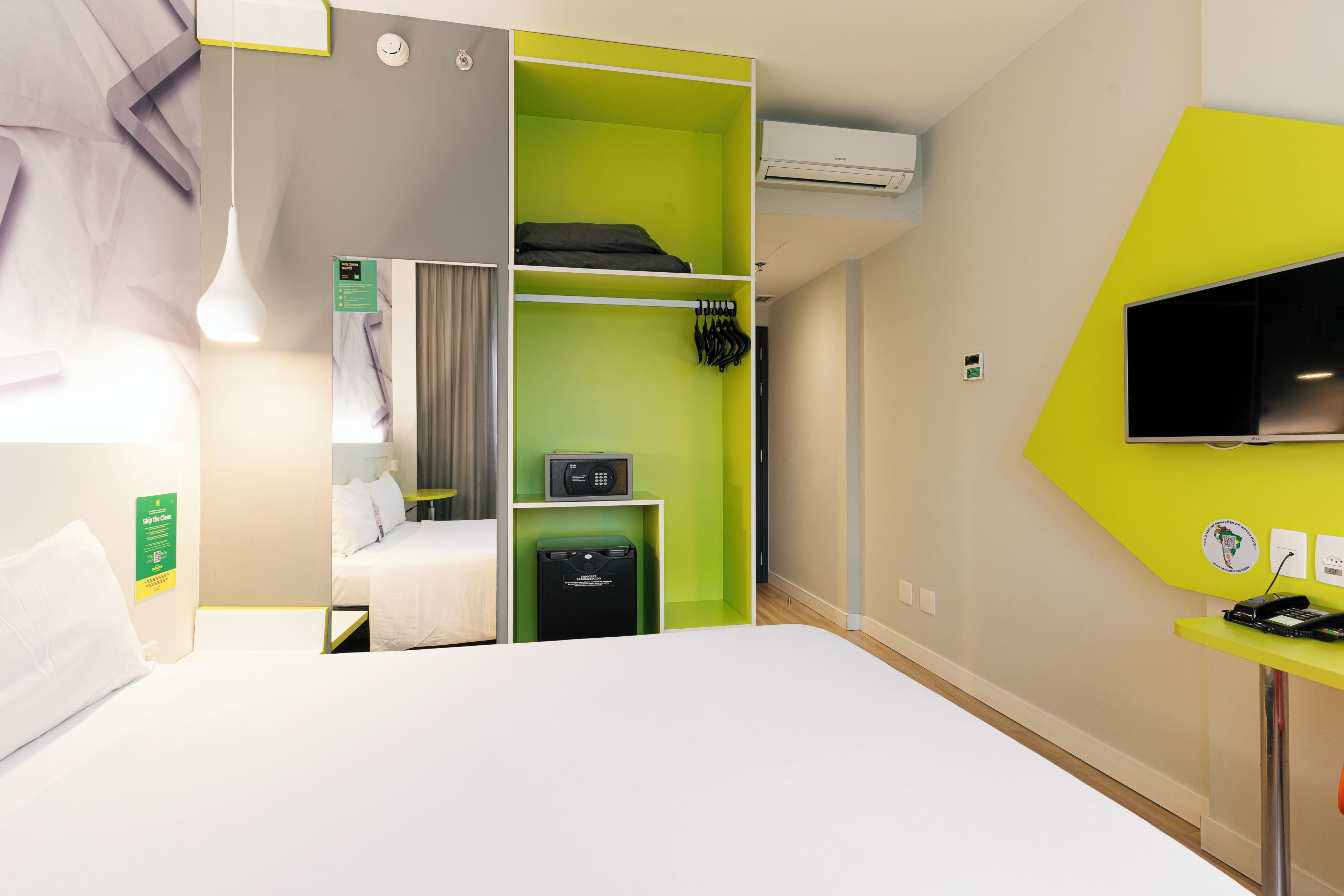 A hotel room with a bed, a green storage unit, a TV, and a desk with a phone.