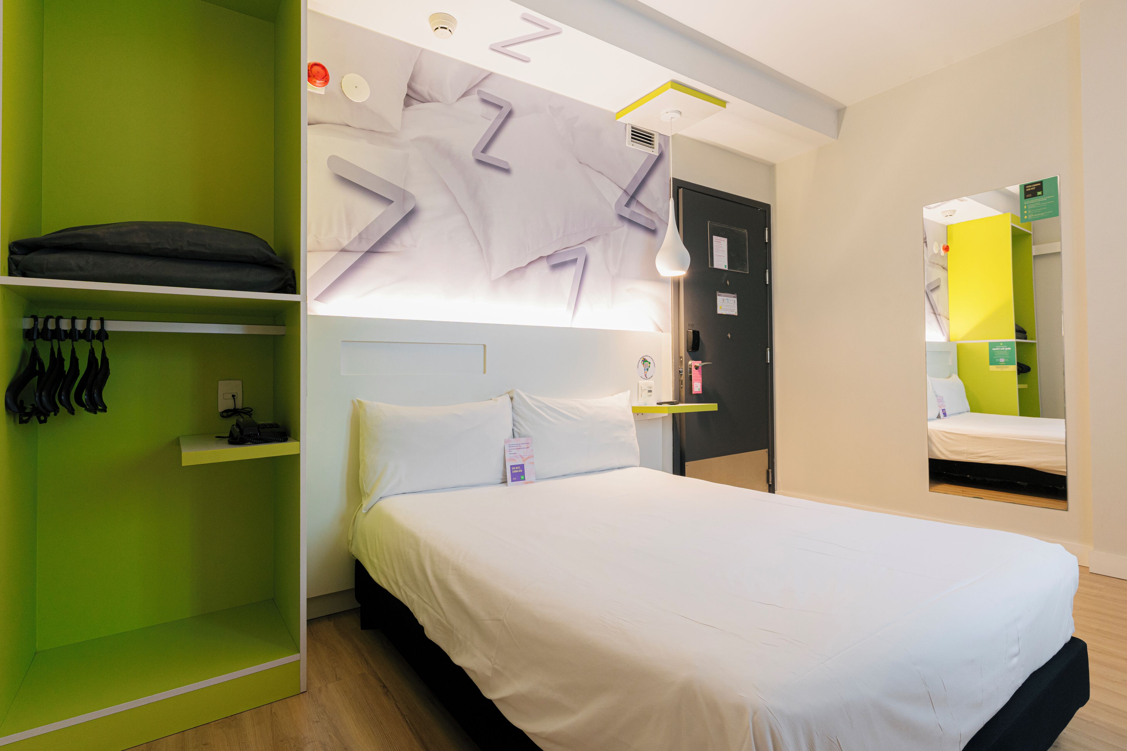 Une chambre d’hôtel moderne avec un lit, une armoire verte et un miroir qui reflète une autre pièce.