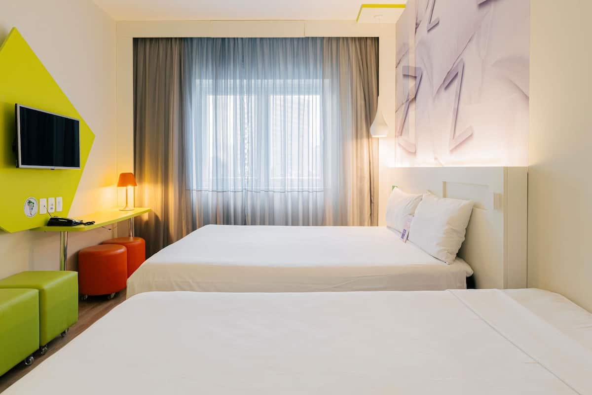 Een hotelkamer met twee bedden, een aan de muur bevestigde televisie, een bureau met een lamp en een groot raam met gordijnen.