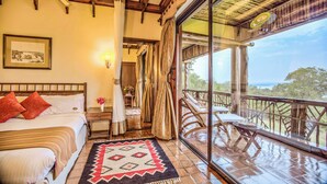 A bedroom with a large bed, a bedside table, a nightstand, a window with curtains, and a balcony with wooden furniture. - Paraa Safari Lodge (Paraa)