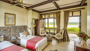A hotel room with two beds, a large window with curtains, a chair, a small table, and a ceiling fan. - Paraa Safari Lodge (Paraa)