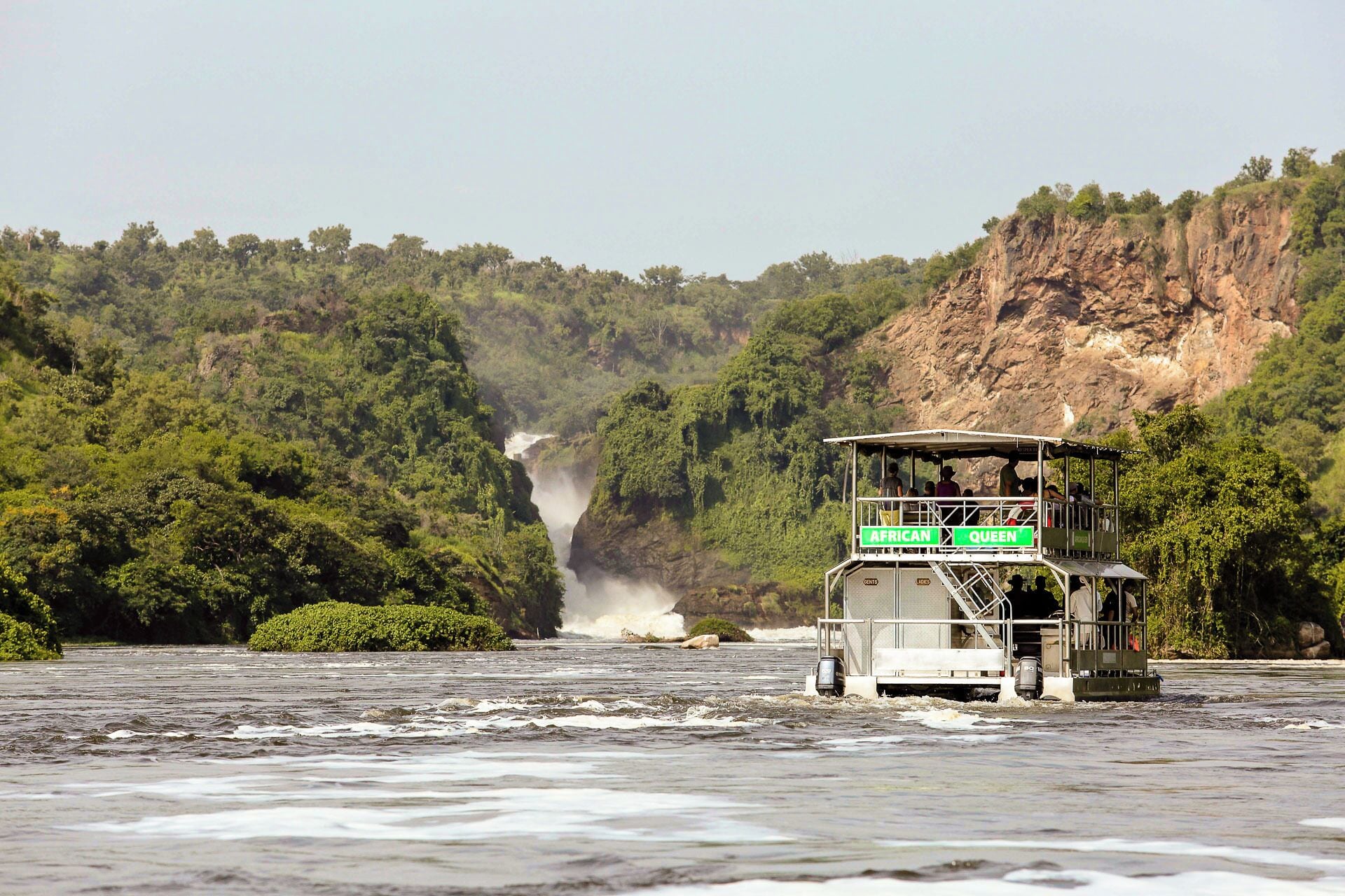 A boat with a viewing platform labeled "African Queen" on a river surrounded by lush greenery and cliffs.