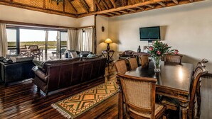 A spacious living room with a large sofa, dining table, and chairs, a ceiling fan, and a TV mounted on the wall.