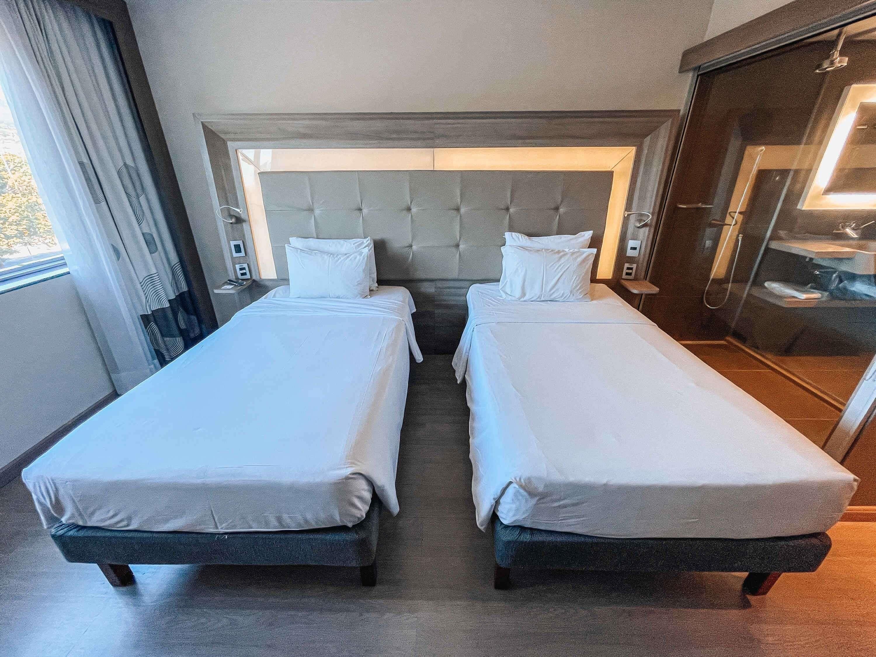 Quarto de hotel com duas camas de solteiro, uma cabeceira grande e acolchoada, área de chuveiro e janela com cortinas.