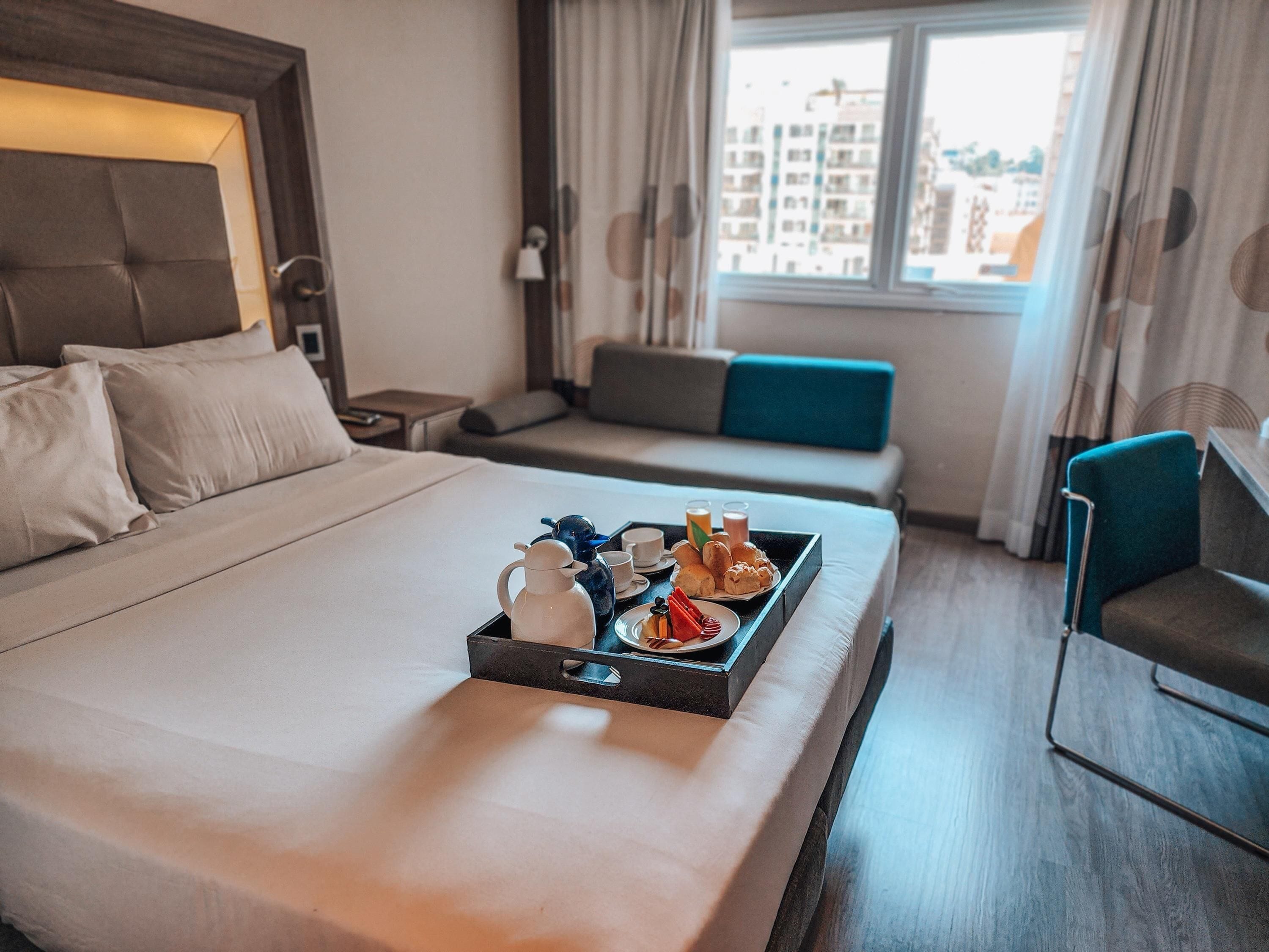 Habitación de hotel con cama, bandeja de desayuno, sofá y vistas a edificios por la ventana.
