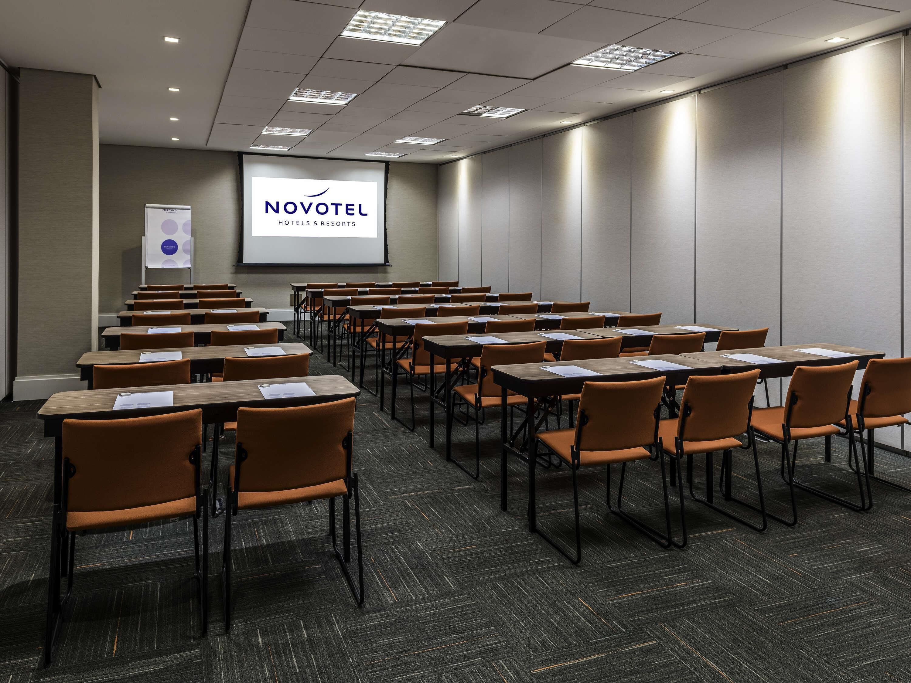 Un salón de conferencias con filas de mesas y sillas, una pantalla de proyección y un cartel de Novotel Hotels & Resorts.