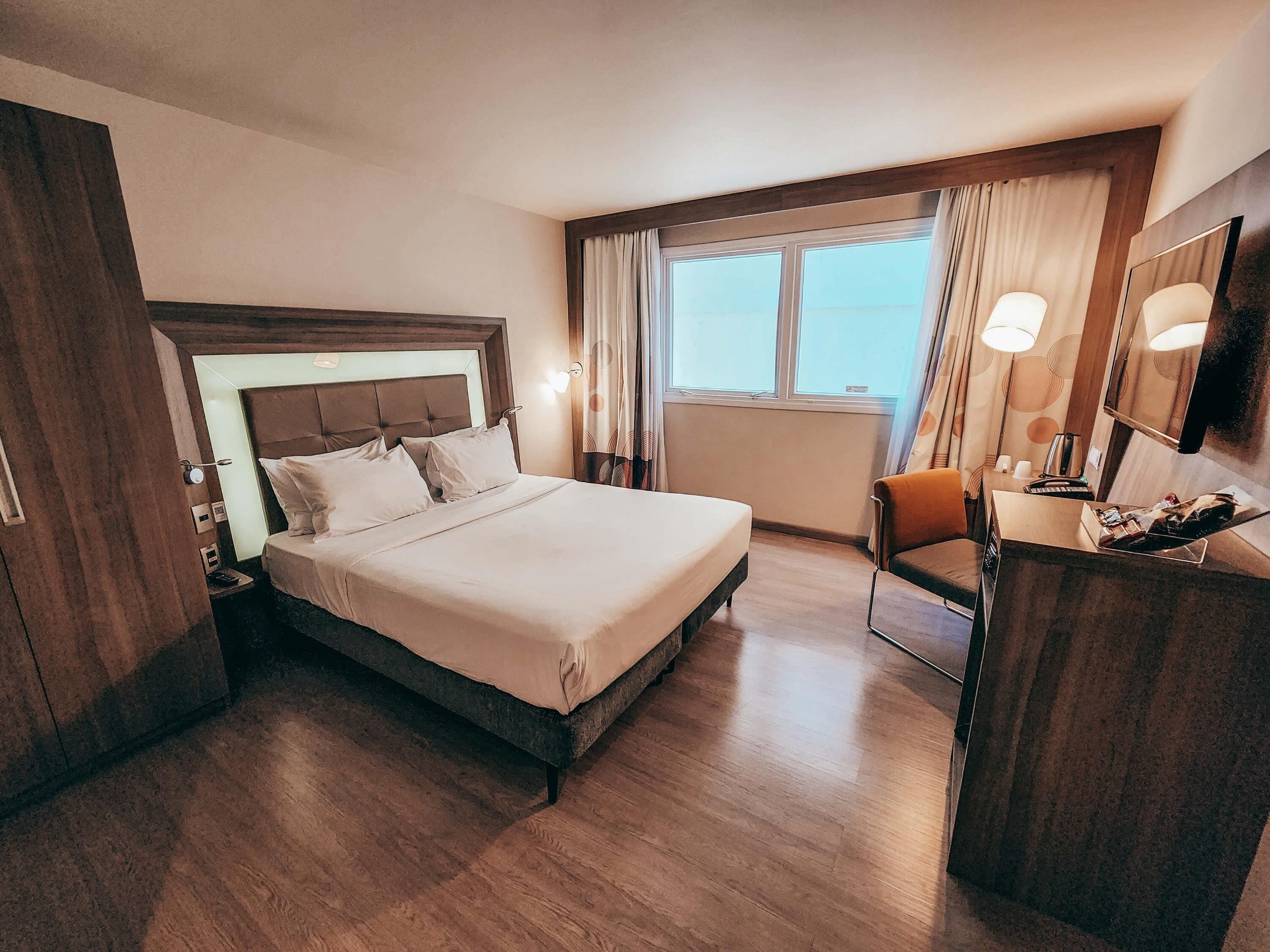 A hotel room with a large bed, a desk with a chair, a television, and a window with curtains.