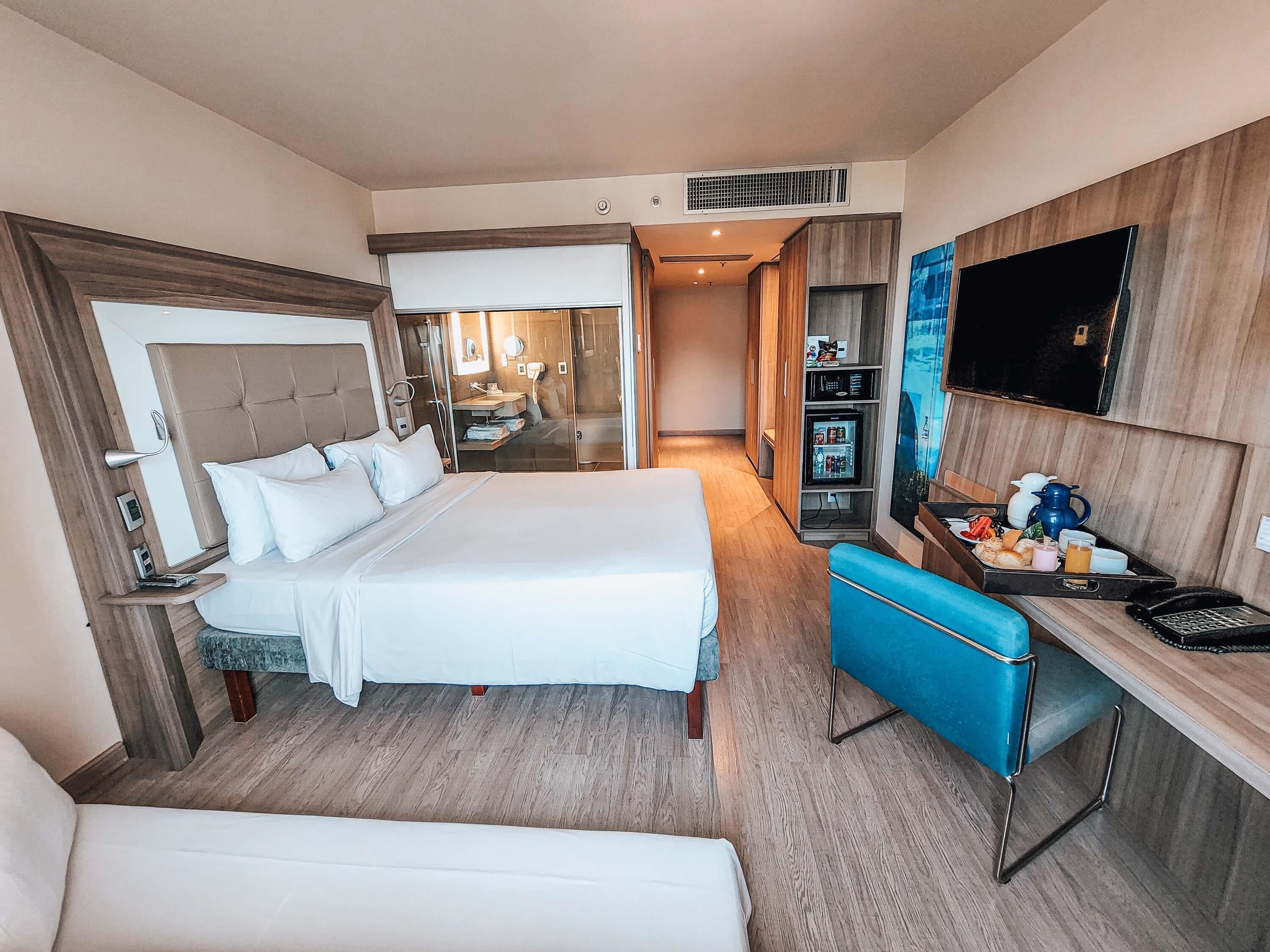 A hotel room with a bed, a desk with a tray of food, a TV, and a minibar.